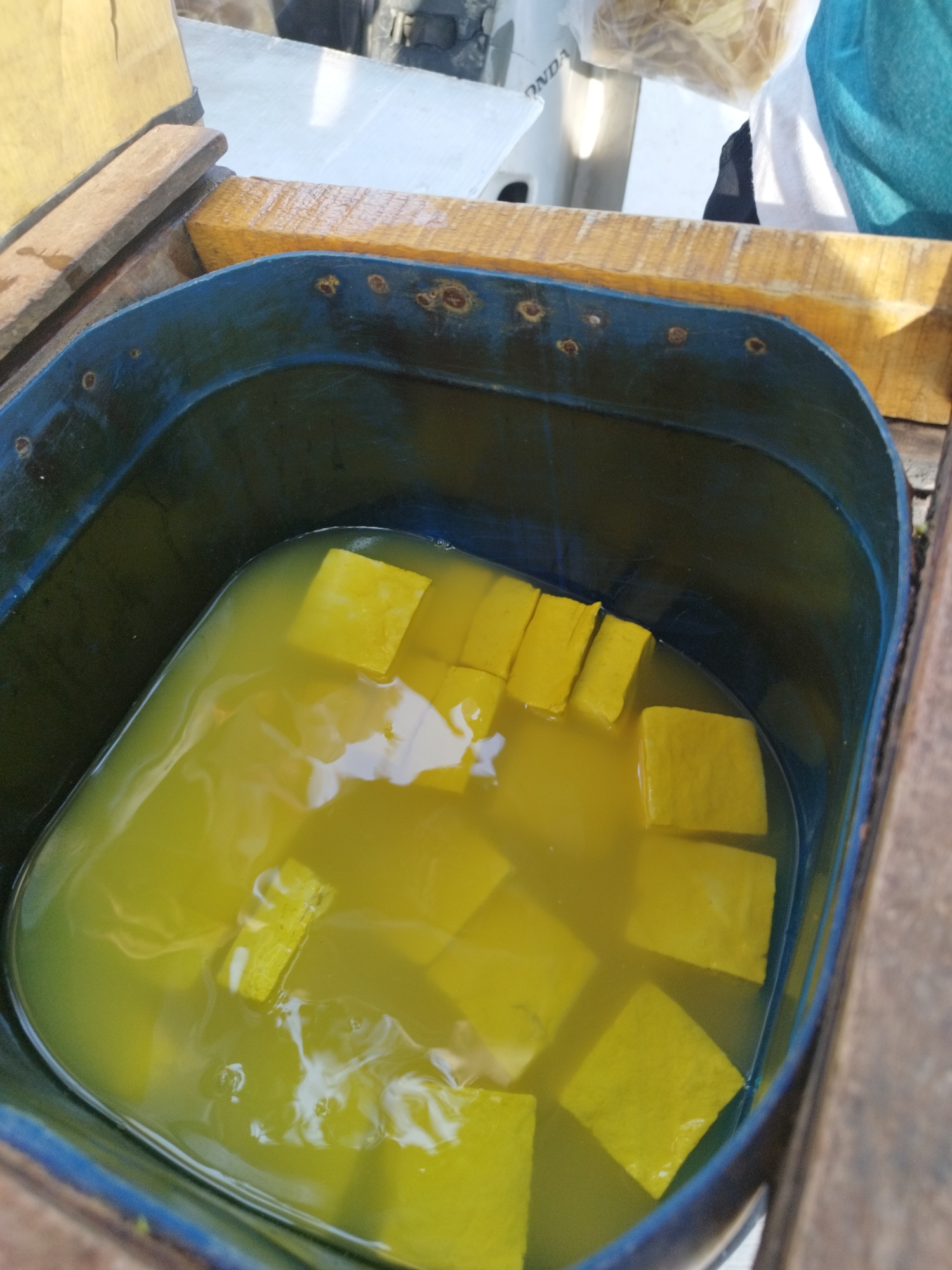 A close-up, high-angle shot shows a blue plastic tub filled with bright yellow liquid, in which numerous yellow, rectangular blocks are submerged. The tub is positioned outdoors, resting on wooden planks. The lighting suggests it is daytime, with sunlight illuminating the scene. The yellow blocks appear to be some form of food product, possibly tofu or a similar item being processed or preserved. The liquid is slightly murky, with some subtle ripples on its surface. The edges of the tub show some wear and tear, with a few rusty spots visible. The overall impression is of a rustic, possibly food-production setting in Karawang, Indonesia.