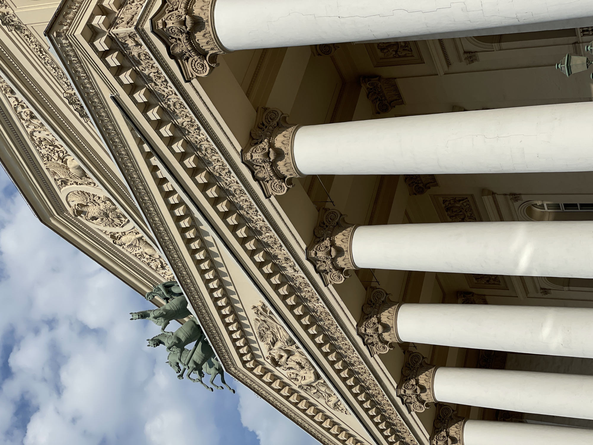 Here's a detailed description of the image:
**Scene:** The image shows a low-angle, upward-tilted view of a section of the Bolshoi Theatre's façade in Moscow, Russia.
**Architecture:** The neoclassical architecture is apparent in the ornate detailing. The architectural elements include:
* **Columns:** Several smooth, white cylindrical columns are visible, supporting the structure above. Each column has an ornate capital.
* **Entablature:** The entablature is heavily decorated with intricate carvings and sculptural elements. There are sections with repeating geometric patterns, as well as sections featuring more organic, floral, or figurative designs.
* **Quadriga:** A partial view of the Bolshoi Theatre's iconic quadriga statue (a chariot drawn by four horses) is visible against a blue sky with light clouds.
**Color and Light:** The color palette is dominated by light beige and white. The architectural details cast shadows due to the angle of the sunlight, creating contrast and highlighting the intricate carvings. The sky in the background is blue with white clouds, providing a contrasting backdrop.
**Overall Impression:** The image emphasizes the grandeur and artistic detail of the Bolshoi Theatre's exterior. The unusual angle provides an interesting perspective on the architecture.
