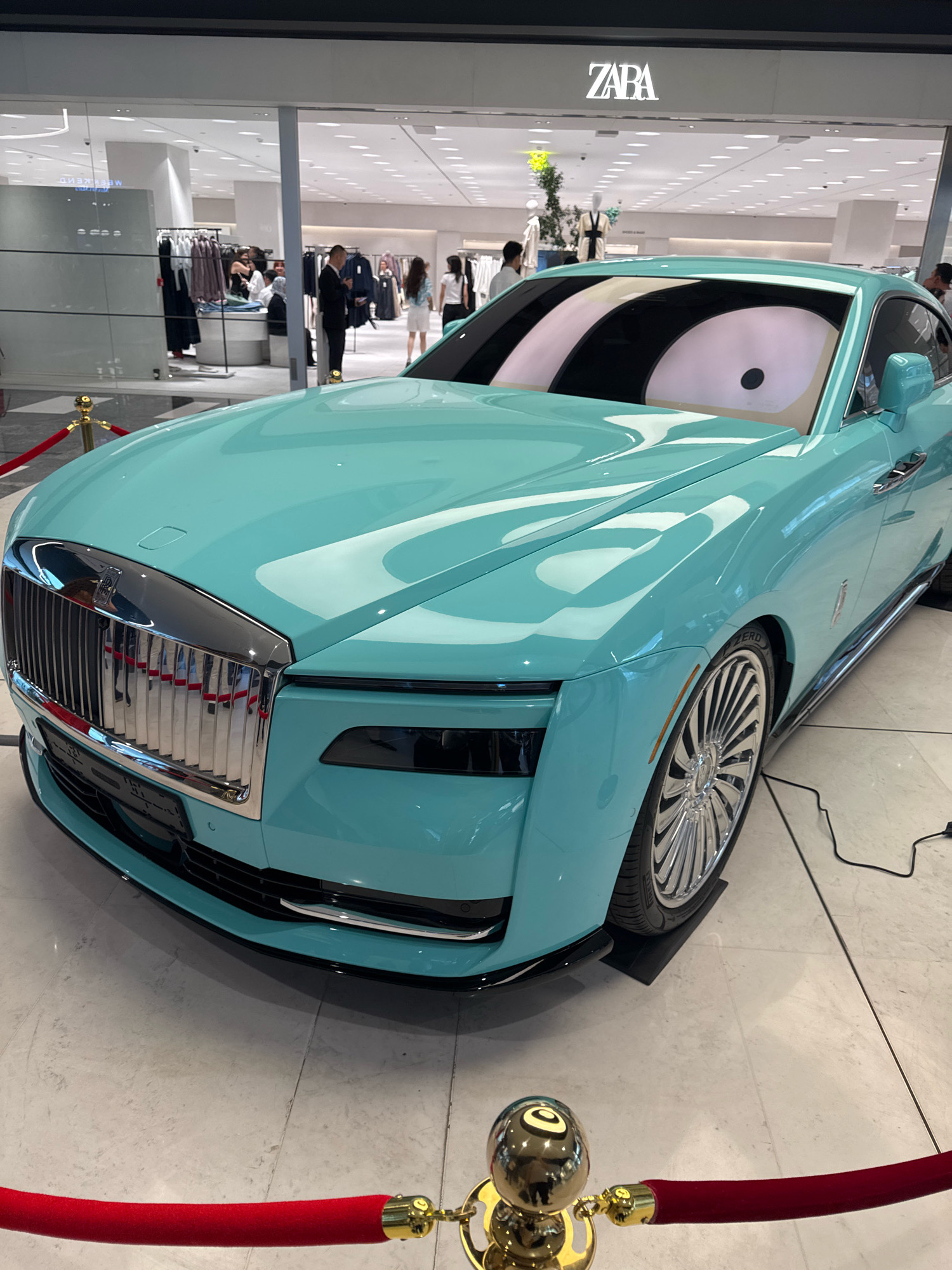 A turquoise Rolls-Royce is parked on a tiled floor inside a modern retail establishment, indicated by the "ZABA" logo visible above. The car's windshield displays a cartoonish eye graphic. Red velvet ropes with gold stanchions cordon off the vehicle. In the background, retail displays with clothing racks and mannequins are visible, along with several shoppers browsing. The environment appears to be a well-lit, upscale shopping mall.
