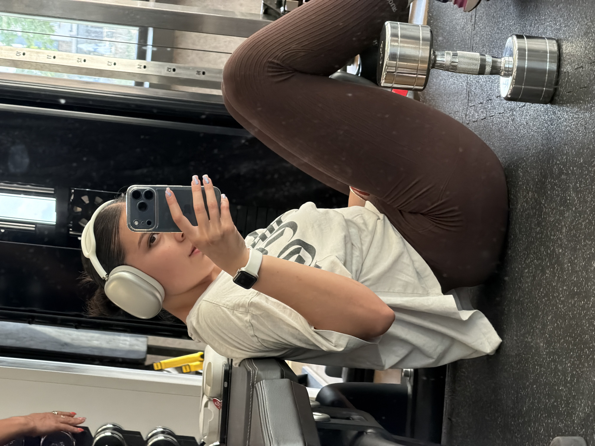 A woman is posing for a selfie in a gym. She is wearing headphones and a white t-shirt, and has brown leggings on. She is using a cell phone with a black case to take the picture. She is leaning on gym equipment. There are dumbbells on the floor next to her. The location is a gym, but the specific city in Bulgaria (Gabrovo) cannot be determined from the image.