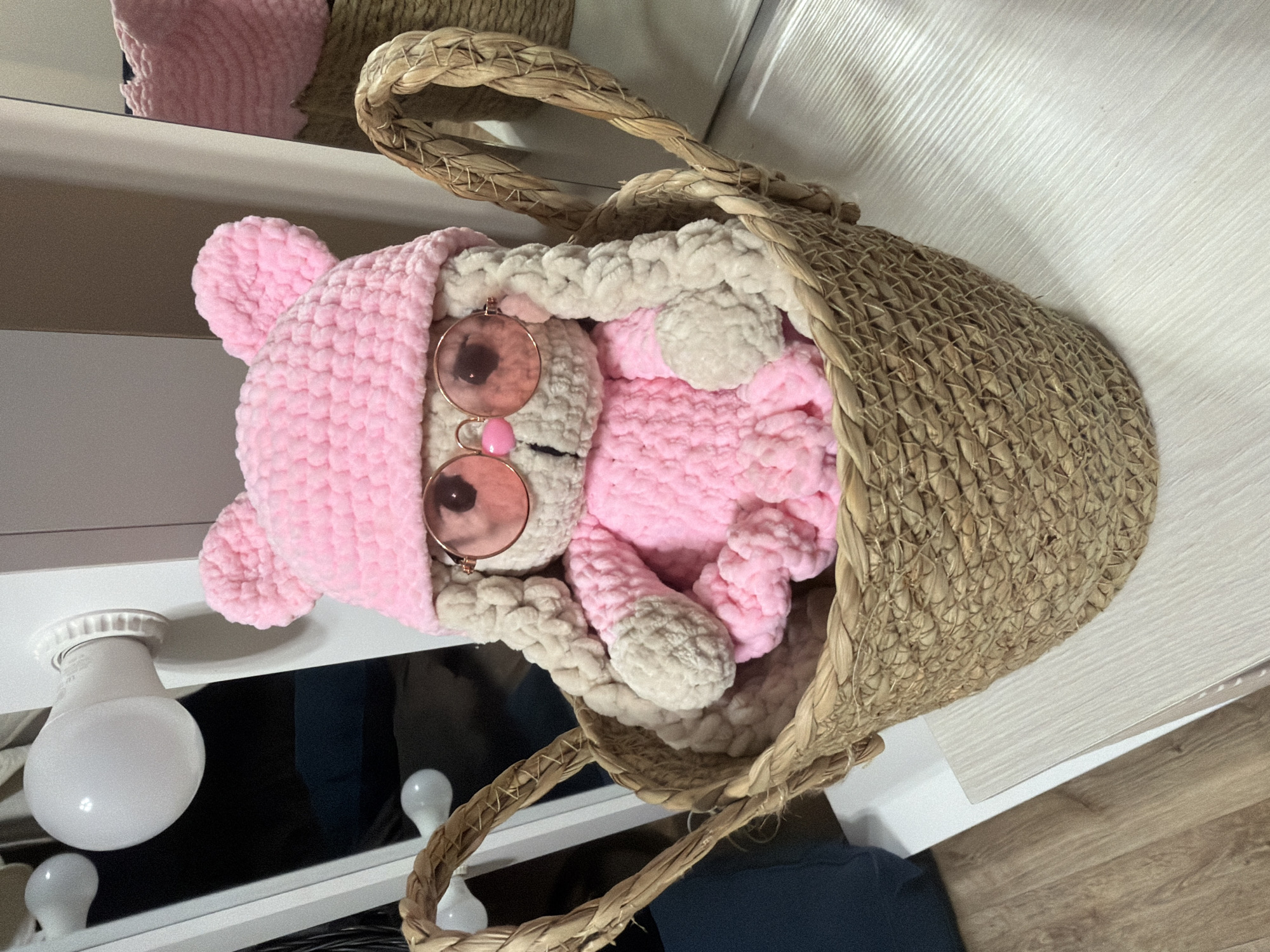 A crocheted stuffed bunny wearing a pink hat with ears and pink sunglasses sits in a woven basket. The bunny has light-colored arms and long, braided ears. Its body is pink and appears to be wearing a ruffled dress. The basket is made of woven straw and has two handles. The bunny is positioned in the center of the basket, which is on a white surface. In the background, there is a white structure with light fixtures and a reflection in a mirror. The environment appears to be indoors, possibly a room in a home or a shop. There is no information in the image to determine its location within the city of Shymkent, Kazakhstan.