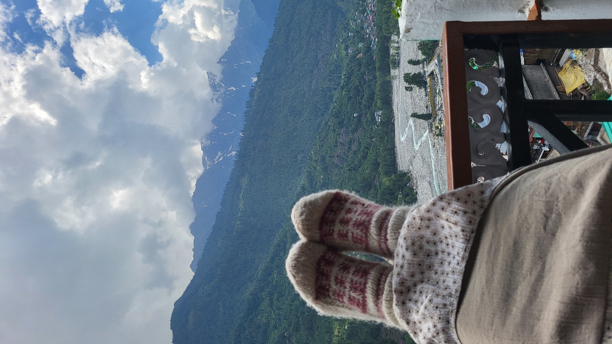 The image displays a first-person perspective looking out from a balcony over a mountainous valley in Vashisht, India. In the immediate foreground, a person's feet, covered in cream-colored knitted wool socks featuring a repeating red and brown patterned design, are visible. The feet rest on a light brown blanket or shawl, which has a cream-colored trim with a subtle, small floral or dotted pattern. Below the feet, a dark wooden balcony railing with a white, carved decorative panel is partially visible on the lower right.
The midground reveals a wide riverbed filled with light-colored rocks and pebbles, through which a narrow, meandering turquoise river flows, consistent with the Beas River. Trees and green vegetation line the riverbanks. Beyond the river, the valley ascends into densely forested hills and mountains, primarily covered with dark green coniferous trees. A cluster of buildings, forming a village, is visible among the trees on the right side of the valley.
In the background, a range of towering mountains with snow-capped peaks extends across the horizon. The sky above is largely covered with a mix of heavy grey and white clouds, with patches of clear blue sky visible predominantly on the right side. The overall scene captures a tranquil, cool environment characteristic of the Himalayan region, specifically the scenic views from Vashisht, a village near Manali in Himachal Pradesh.