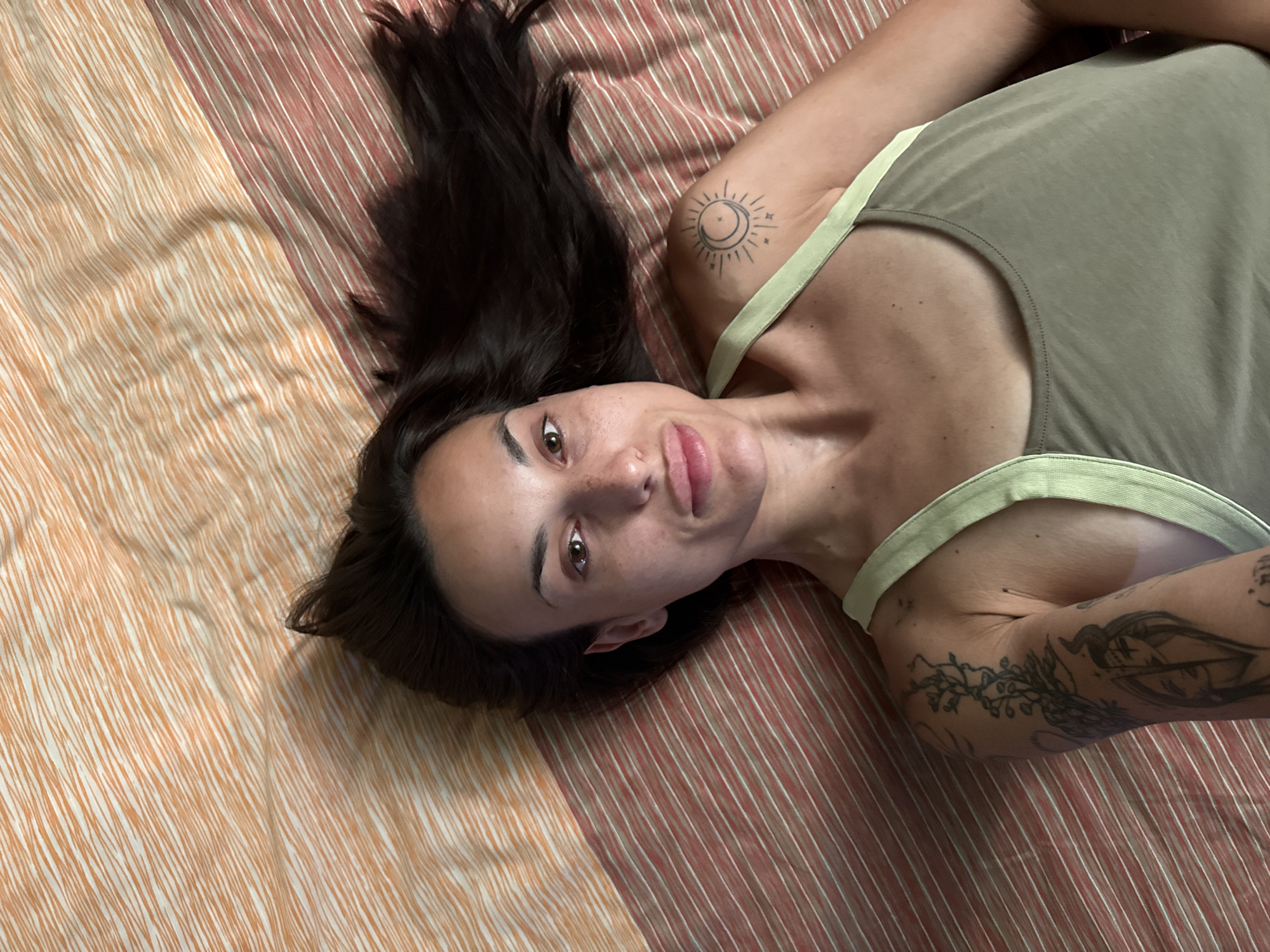 A young woman with dark hair and fair skin is lying on a bed with striped linens. She is wearing a light green tank top and has several tattoos on her arms, including a sun and moon design on her shoulder and a botanical design on her upper arm. The image appears to have been taken from a high angle, looking down at the woman. The bed linens are striped in shades of orange and white, and red and white. The lighting is soft and natural, suggesting the photo was taken indoors during the daytime. There is no indication of the specific location within the city of Pleven, Bulgaria.