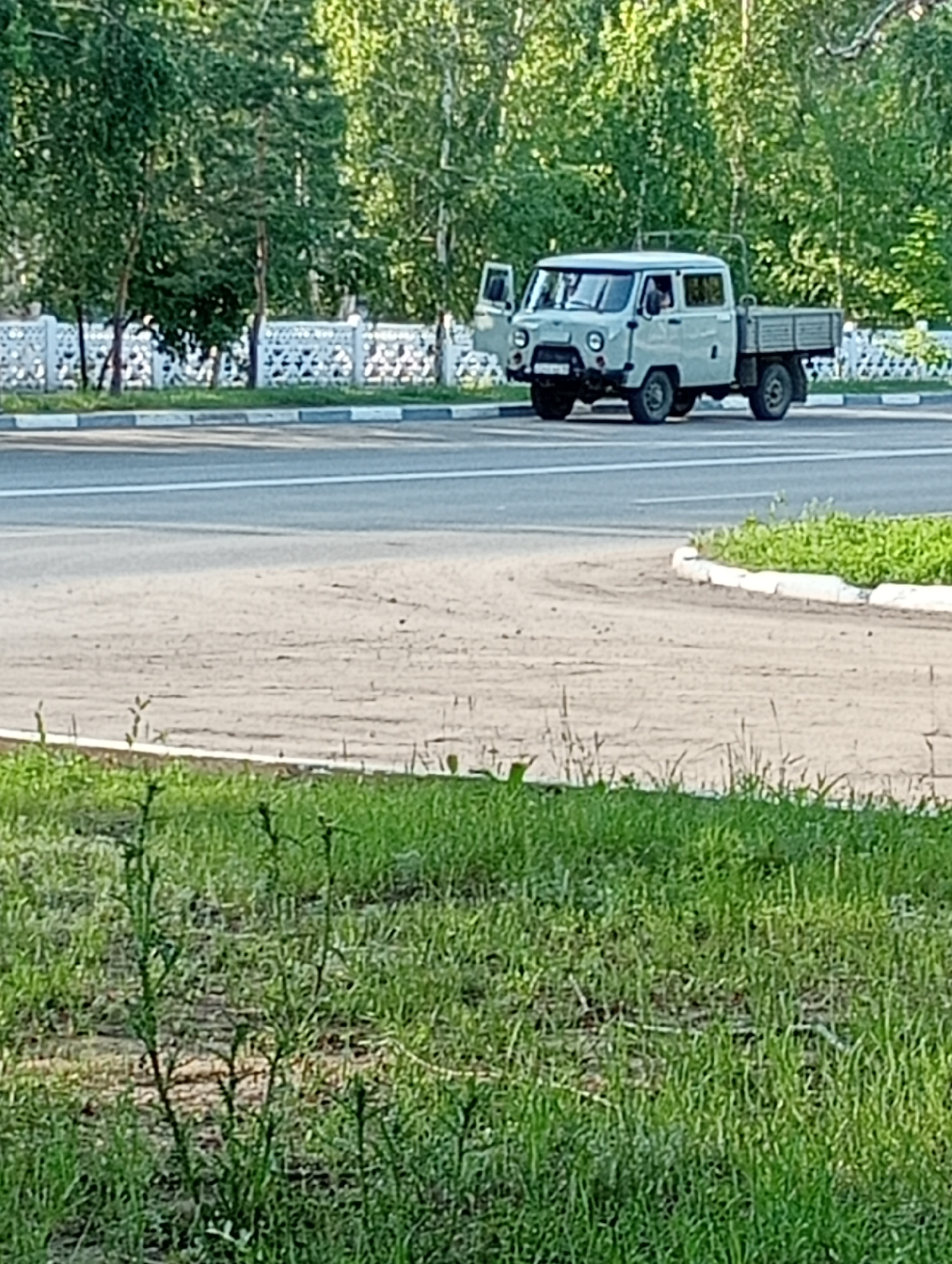 A light-colored, four-door UAZ pickup truck is parked on a dirt lot near a paved road in Kostanay, Kazakhstan. The truck's driver-side door is open, and a person is visible in the driver's seat. The truck has a flatbed in the rear. A white decorative fence runs along the side of the road, and a line of trees is visible behind the fence. The foreground of the image consists of grass.