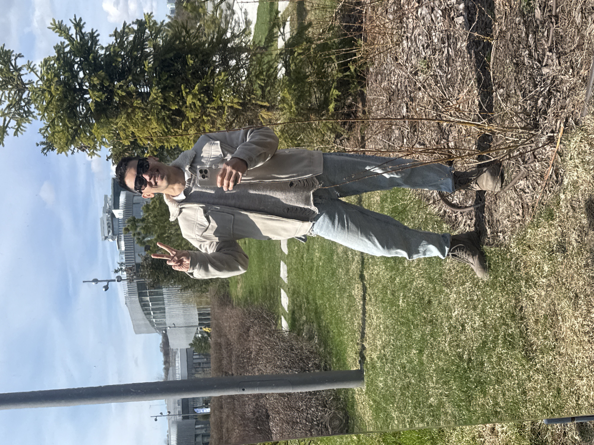 The image displays a male individual posing outdoors in Kazan, Russia, on a bright, partly cloudy day. The subject, an adult man, is smiling directly at the camera, making a "peace" sign with his left hand. He wears sunglasses and holds a light-colored smartphone in his right hand. His attire includes a layered light-colored jacket with a shearling-like collar and lining, over a darker top, paired with light blue distressed jeans and dark boots.
The setting appears to be an urban green space or park. The man stands on a grassy area, with dried leaves and sparse brown brush visible behind him to his right. To his left and further back, several vibrant green conifer trees are prominent. In the background, modern buildings with reflective facades are visible under a blue sky with white clouds. A dark grey pole and a reflective surface, possibly a barrier or wet ground, are in the lower foreground. The man's expression and pose convey a cheerful and relaxed demeanor. No discernible text is visible in the image.