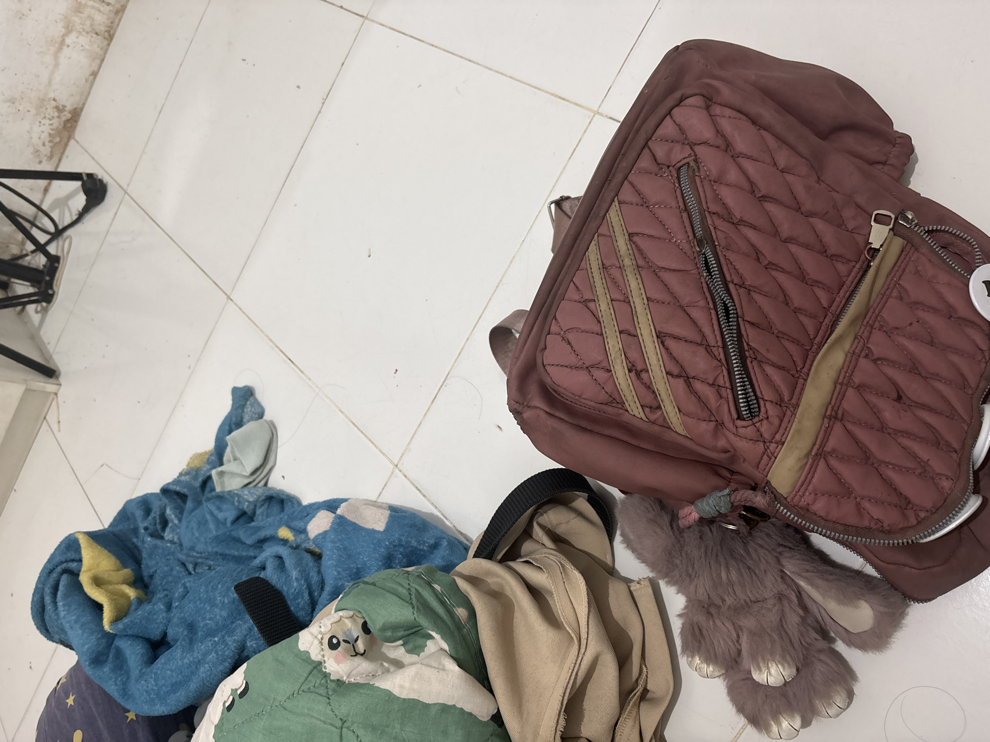 The image depicts a cluttered scene on a white tiled floor, likely inside a residential or storage area in North Jakarta, Indonesia. The foreground is dominated by a rumpled pile of items including a rose-colored quilted backpack with tan accents and zippers, a plush gray toy with floppy ears and paws, and a folded tan garment. To the left, a green quilted item with a cartoonish face and a blue blanket with yellow accents are visible. Some small, light blue shoes are also present in the background. The environment appears unkempt, with the floor showing signs of dust and dirt, and a section of a wall with water damage is visible in the upper right corner. A black metal object, possibly part of a stand or furniture, is also partially visible in the background. There are no people or observable actions taking place in the image.