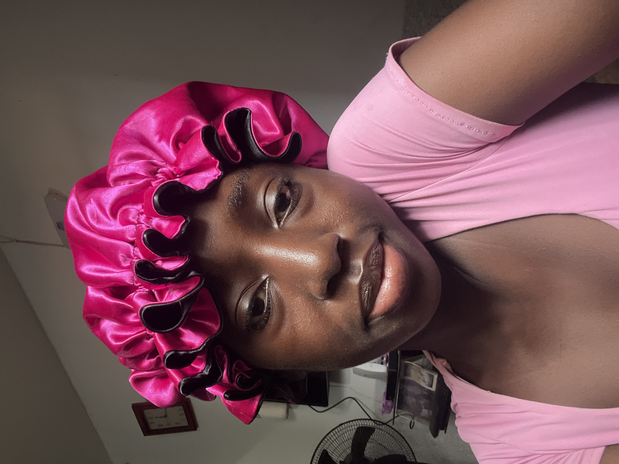 A medium shot depicts a dark-skinned woman wearing a pink shirt and a pink satin bonnet with black trim. Her face is angled toward the camera. The background contains a wall, a fan, and various objects on a shelf or table. No specific information indicates the image was taken in Abuja, Nigeria.