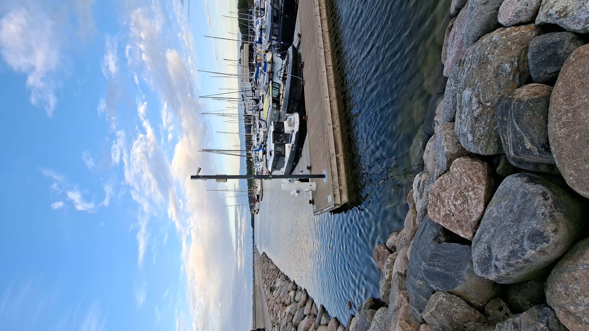 The image depicts a view of a marina in Tallinn, Estonia, likely the Pirita harbor, given the distinctive breakwater and the general scenery. The foreground is dominated by a rocky breakwater made of large, weathered stones, creating a natural, rugged barrier. Beyond the breakwater, a calm body of water reflects the sky, which is a mix of blue with scattered, soft white clouds, indicating either early morning or late afternoon light.
The water leads into the main marina area where numerous sailboats and motorboats are docked. The boats vary in size and type, with masts of sailboats creating a distinct silhouette against the horizon. Several motorboats are moored to a wooden dock in the mid-ground, one of which is a black and white vessel with visible registration numbers. A security camera is mounted on a pole on this dock.
On the left side of the image, a person can be seen walking along the paved walkway on top of the stone breakwater. The background shows more boats and the distant shoreline with some buildings, suggesting the urban edge of the city. The overall atmosphere is serene and peaceful, capturing a typical day at a Baltic Sea marina.