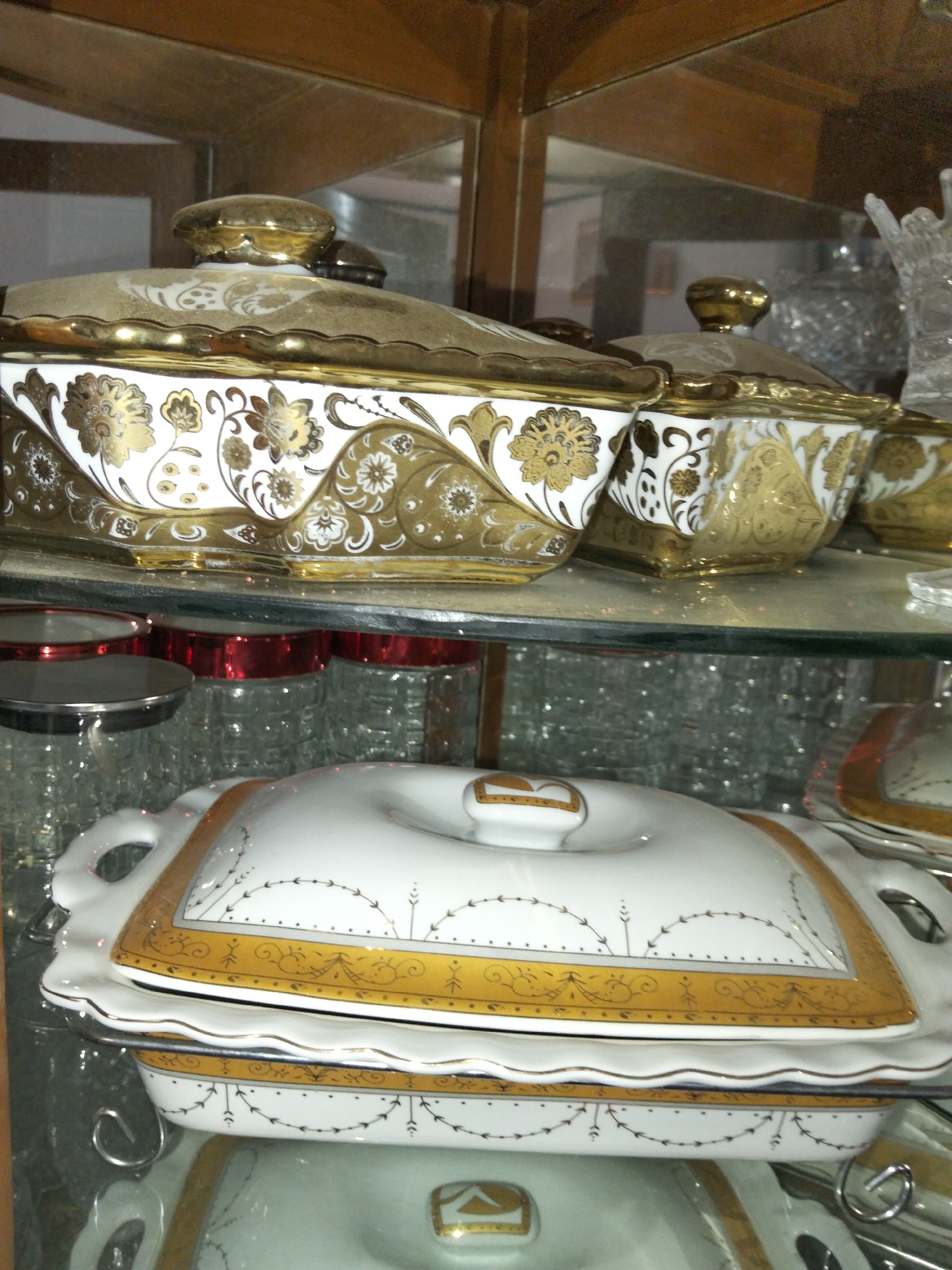 The image shows a display cabinet with several pieces of fine china, predominantly white with intricate gold detailing. On the top shelf, there are at least two serving dishes with lids. The dish on the left is larger and appears to be an oval-shaped tureen with a scalloped edge and a gold floral pattern. The lid has a gold knob. To the right of it, another similar serving dish is visible, also with gold detailing.
On the shelf below, a white serving dish with a lid is prominently displayed. This dish has a rectangular shape and features a gold decorative band with a delicate scroll pattern and small arrow-like motifs around its perimeter. The lid also has a gold knob. Beneath this dish, another white serving dish is partially visible.
In the background, on the top shelf, a large, clear, cut-glass object, possibly a vase or decanter, is present. The cabinet itself appears to be made of dark wood with glass shelves. The overall impression is one of elegance and a display of decorative tableware. The scene is located within a home or retail environment. There are no people or animals present in the image.
