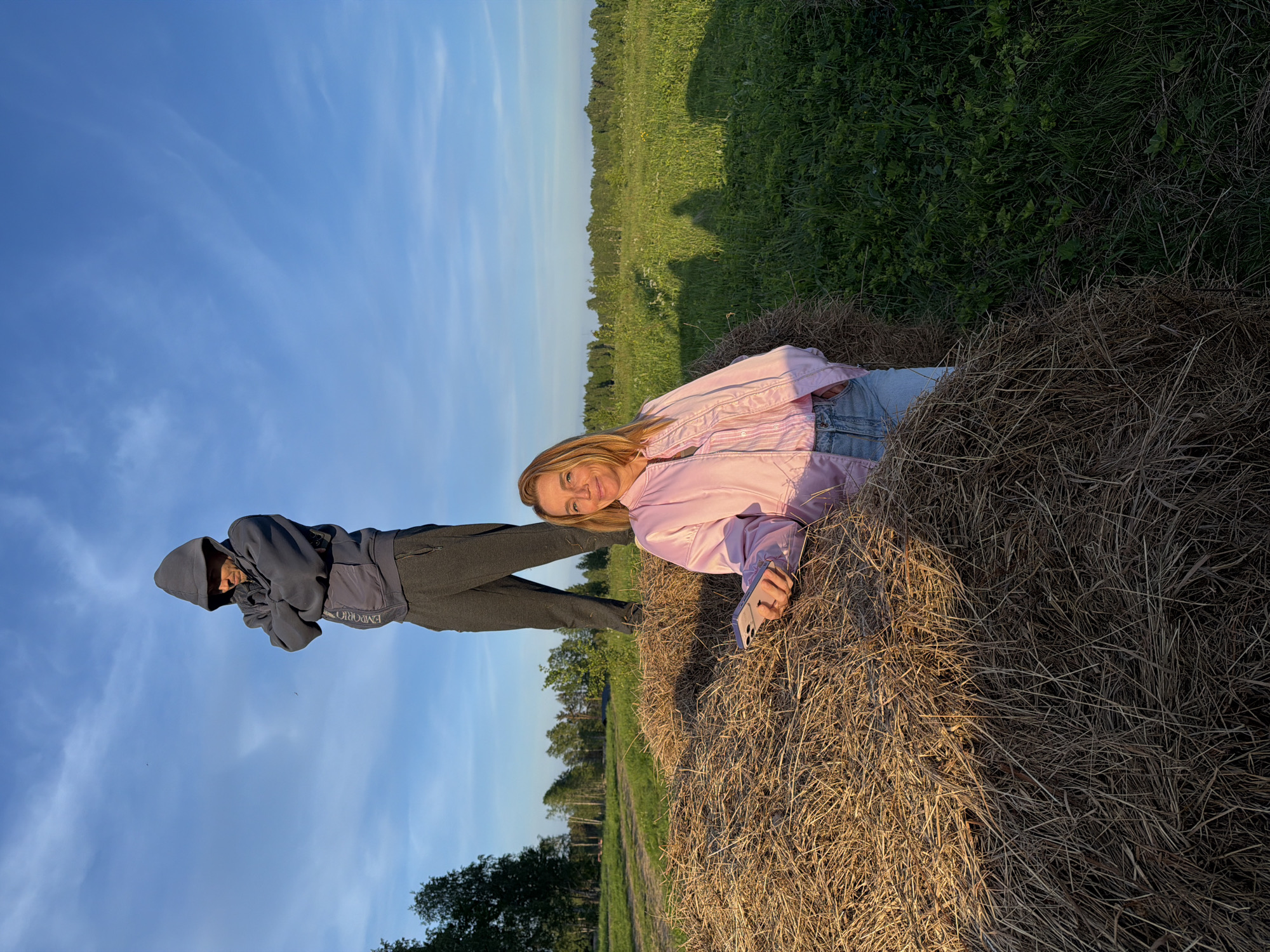 This image depicts two individuals in an expansive, sunny rural landscape, likely in Vaskelovo, Russia, during the golden hour of late afternoon or early evening.
On the right side of the frame, a person with blonde hair, wearing a light pink jacket and blue jeans, is comfortably reclining on a large, dried hay bale. This individual is smiling pleasantly towards the camera and is holding a light-colored smartphone. To the left of the hay bale, another person stands, dressed in a dark grey hooded sweatshirt and dark pants. Their face is partially obscured by the hood, but they appear to be looking in the direction of the camera or the other person. The text "EMPORIO" is visible on the front of their sweatshirt.
The background features a vast green field that stretches to a distant treeline under a bright blue sky with scattered, wispy white clouds. The warm, directional sunlight illuminates the subjects and the hay bale, creating long shadows across the grass. The overall atmosphere is tranquil and relaxed, capturing a pleasant moment in a natural, open-air setting.