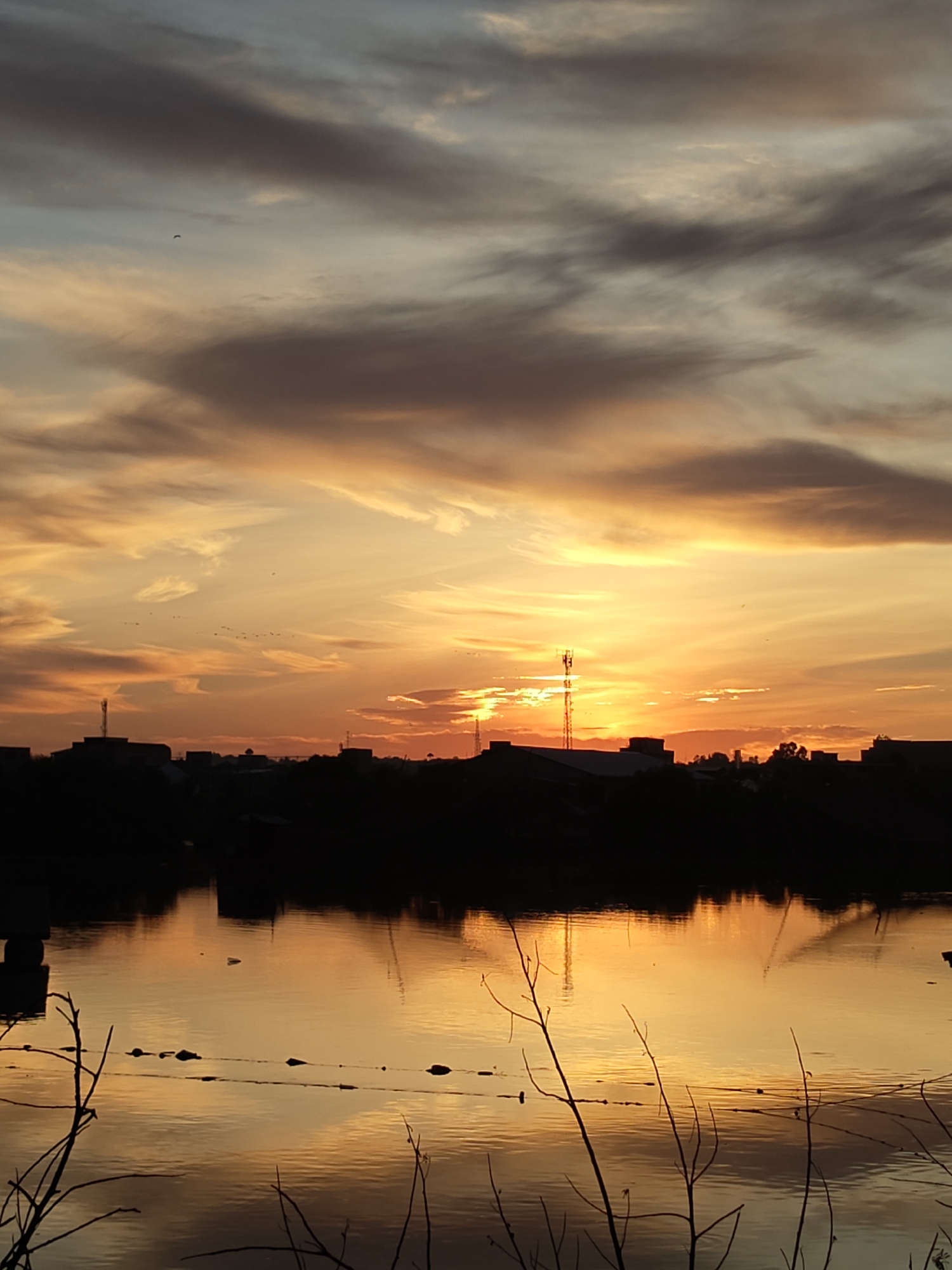 The image captures a sunset over a body of water, likely a river or lake, in Lhokseumawe, Indonesia. The sky is ablaze with vibrant orange and yellow hues, interspersed with darker, dramatic clouds. Silhouetted against this fiery backdrop is the city skyline, featuring buildings and communication towers. The water reflects the colors of the sunset, creating a shimmering, golden surface. Bare tree branches in the foreground add a stark contrast to the luminous sky and water. The scene is serene, with a sense of calm as day transitions into night.