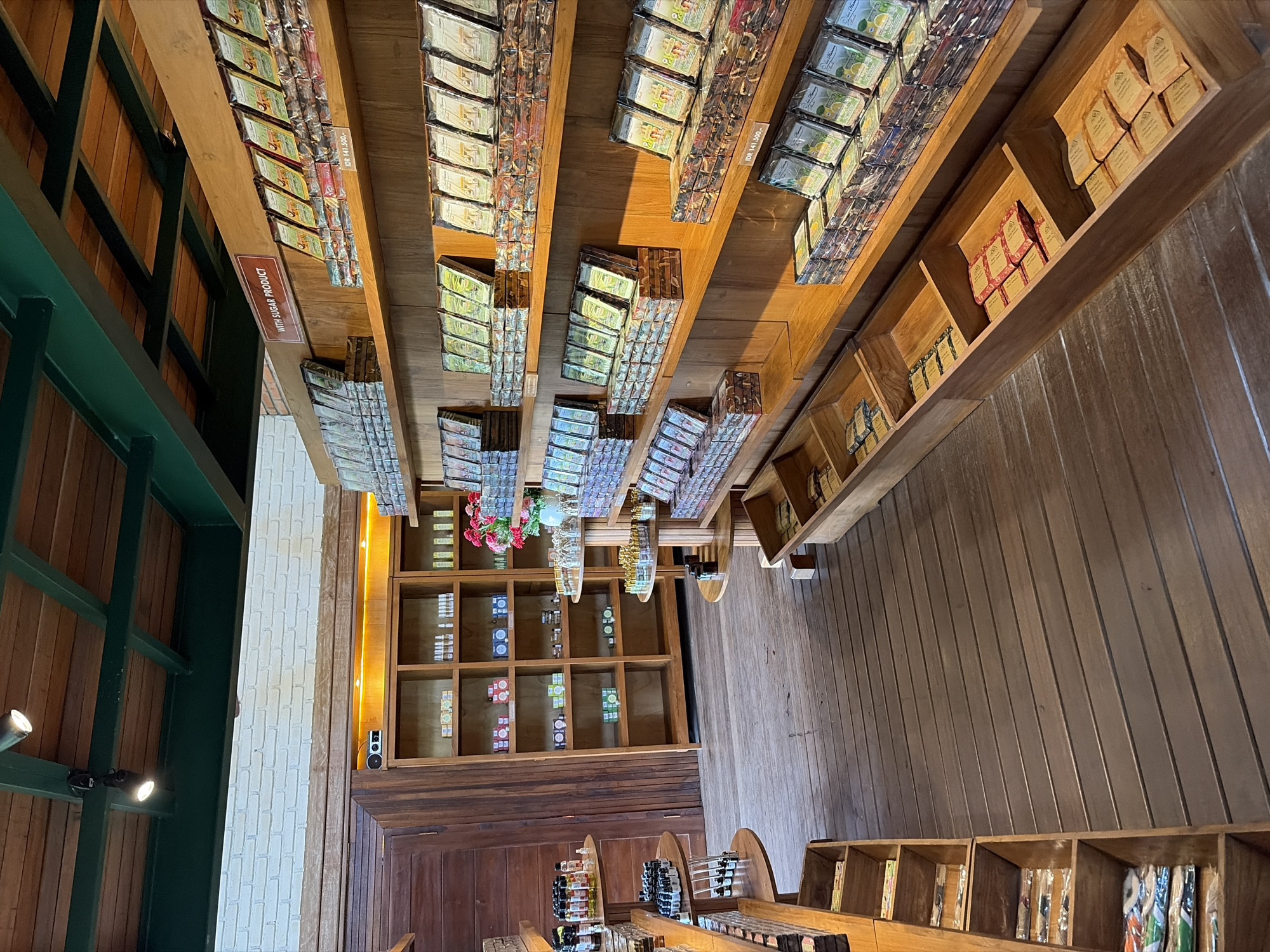 The image depicts the interior of a retail establishment, likely a gift shop or specialty store, in Tegallalang, Indonesia. The store features an abundance of wooden shelving units filled with various products, primarily packaged goods and small bottles or containers.
Dominating the scene are tall, narrow wooden shelves on the right side of the image, displaying rows of stacked products, many of which appear to be chocolate bars or similar confections. Some shelves have labels indicating "WITH SUGAR PRODUCT" and prices in Indonesian Rupiah. In the center background, a larger set of open wooden shelving units are stocked with smaller items, some in colorful boxes and others appearing to be bottles. A vase with artificial flowers is placed on one of these shelves.
To the left, a series of smaller, tiered circular wooden display tables are laden with various products, including bottles and small packages. Another vertical wooden shelving unit on the far left also holds stacked items. The floor is constructed of dark wooden planks. The overall aesthetic is natural and rustic, with extensive use of wood throughout the store's design and displays. No people are visible in the image. The lighting appears to be a mix of overhead fixtures and ambient light, creating a warm glow. The store's ceiling is also wooden, with exposed beams.