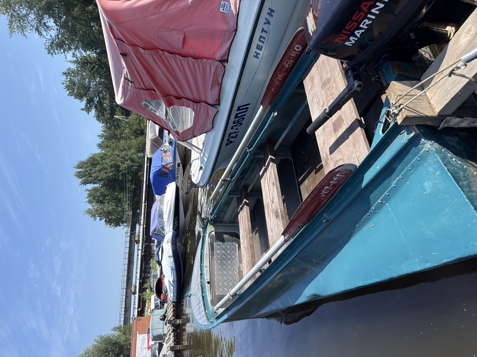 The image shows a marina with several boats docked on a body of water. In the foreground, a teal-colored boat with wooden seats and oars is partially visible. Behind it, a white boat with a red canopy is docked, featuring Russian text and registration numbers. Other boats, some with blue covers, are visible further back. A bridge and trees line the background, suggesting an outdoor setting in Chaikovsky, Russia. The sky is clear and blue, indicating a sunny day.