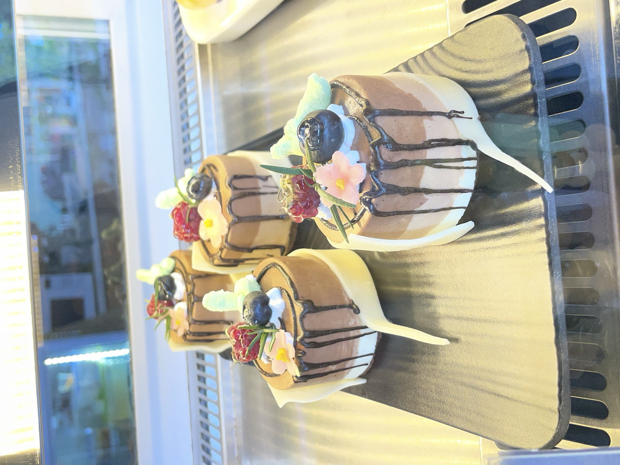 Four individual, elaborately decorated desserts are displayed in a glass case, likely within a bakery or cafe in tp. Đà Lạt, Vietnam. The desserts are cylindrical, resembling mini log cakes or mousse rolls, and are arranged diagonally on a black, textured serving stand with ridged details.
Each dessert features three distinct horizontal layers in shades of brown and off-white, suggesting different flavors like dark chocolate, milk chocolate, and vanilla or white chocolate mousse. They are adorned with dark chocolate drizzle stripes across their tops and sides.
The top of each cake is garnished with a variety of toppings: a single dark berry (possibly a blueberry), a cluster of small red berries (likely raspberries), a delicate pink or light-colored sugar flower, and small green sprigs that resemble rosemary or mint leaves. A small, light green, fluffy-looking candy or sugar decoration (perhaps a marshmallow or meringue piece) and a dollop of white cream or frosting are also visible. A prominent white, pointed, curved chocolate piece is inserted vertically into one end of each cake, adding an elegant flourish.
The display case creates reflections on the glass panel to the left, showing blurred outlines of objects and light sources in the background. The interior of the display case has metallic elements, including a grated shelf or cooling unit on the right. The lighting is bright, illuminating the desserts clearly. No text is visible in the image.