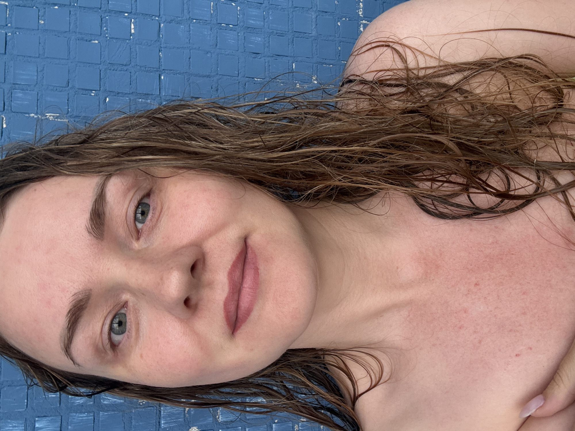 A woman with wet, light brown hair is pictured outdoors against a blue tiled wall. She has fair skin with some redness on her chest and neck. Her eyes are green, and she is wearing a light pink lipstick. The scene is consistent with an outdoor setting in Chaikovsky, Russia, where blue tiled walls are common in public spaces.