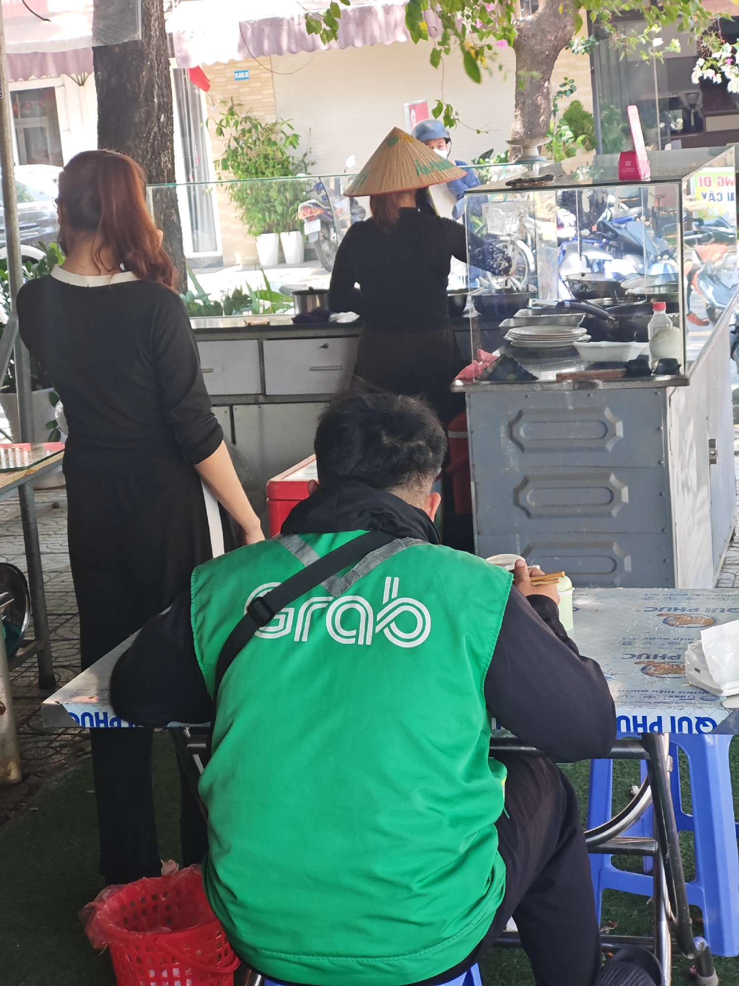 This image depicts an outdoor food stall in Da Nang, Vietnam. A person wearing a green Grab delivery vest is seated at a table, holding chopsticks and a cup. Another person, a woman with red hair, stands facing away from the camera, looking towards the food stall. Behind her, another person in a conical hat is visible near the food preparation area. The stall features a glass display case with cooking utensils and dishes. The ground appears to be covered in artificial turf, and a red plastic basket is visible near the seated person's feet. The scene is set against a backdrop of buildings and vegetation, suggesting an urban environment.
