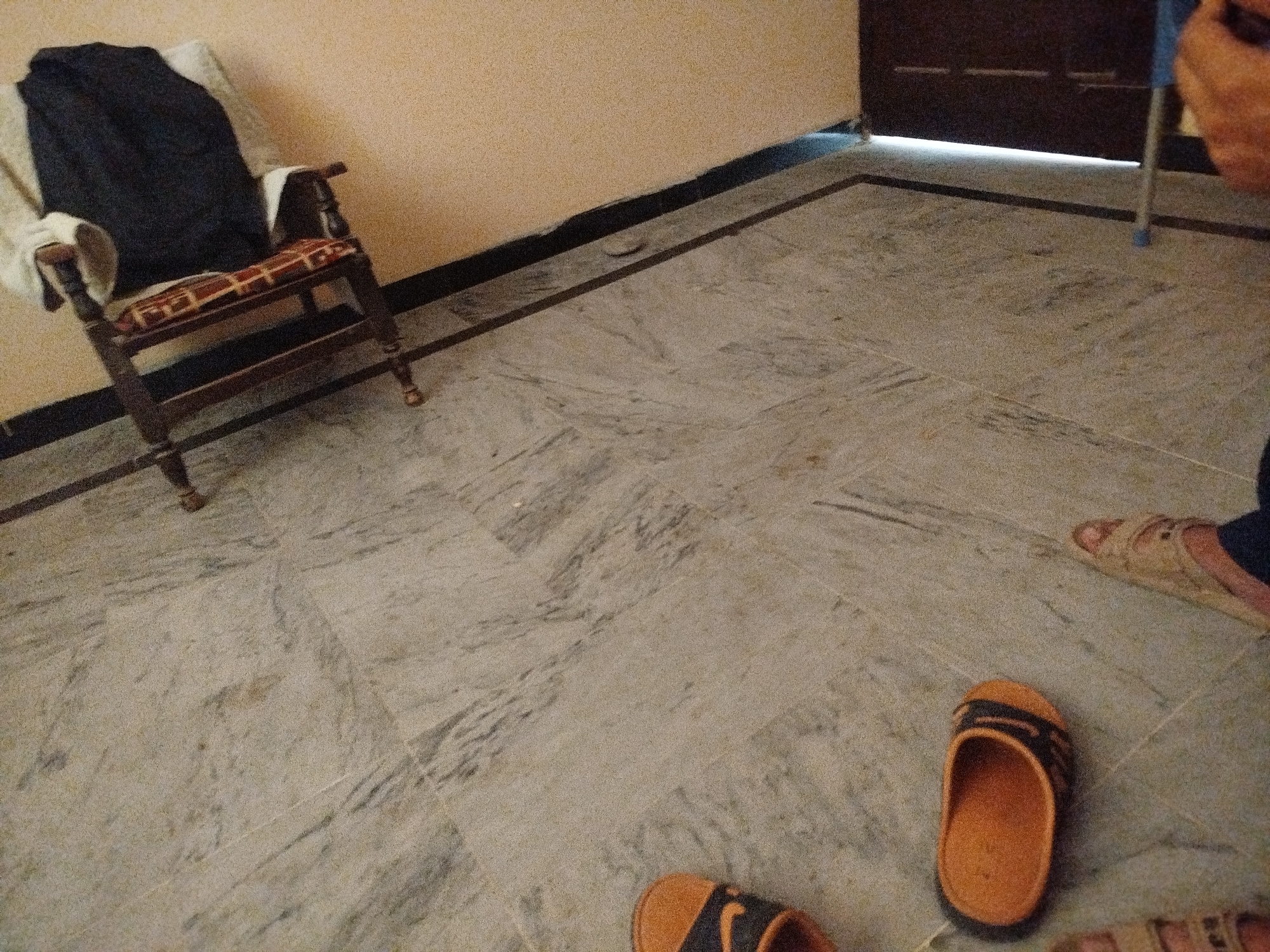The image depicts an indoor scene with marble flooring and a light-colored wall. A wooden chair with a patterned seat and armrests is positioned against the wall, with a dark garment draped over its back. Near the chair, two pairs of slippers are visible on the floor. One pair is orange with black straps, and another is light-colored and worn by a person whose legs and feet are partially visible, wearing dark trousers. The background includes a dark wooden door or cabinet. The lighting suggests an indoor environment, possibly a residential setting. The image does not provide specific clues to pinpoint the exact location within Lassan, Pakistan.
