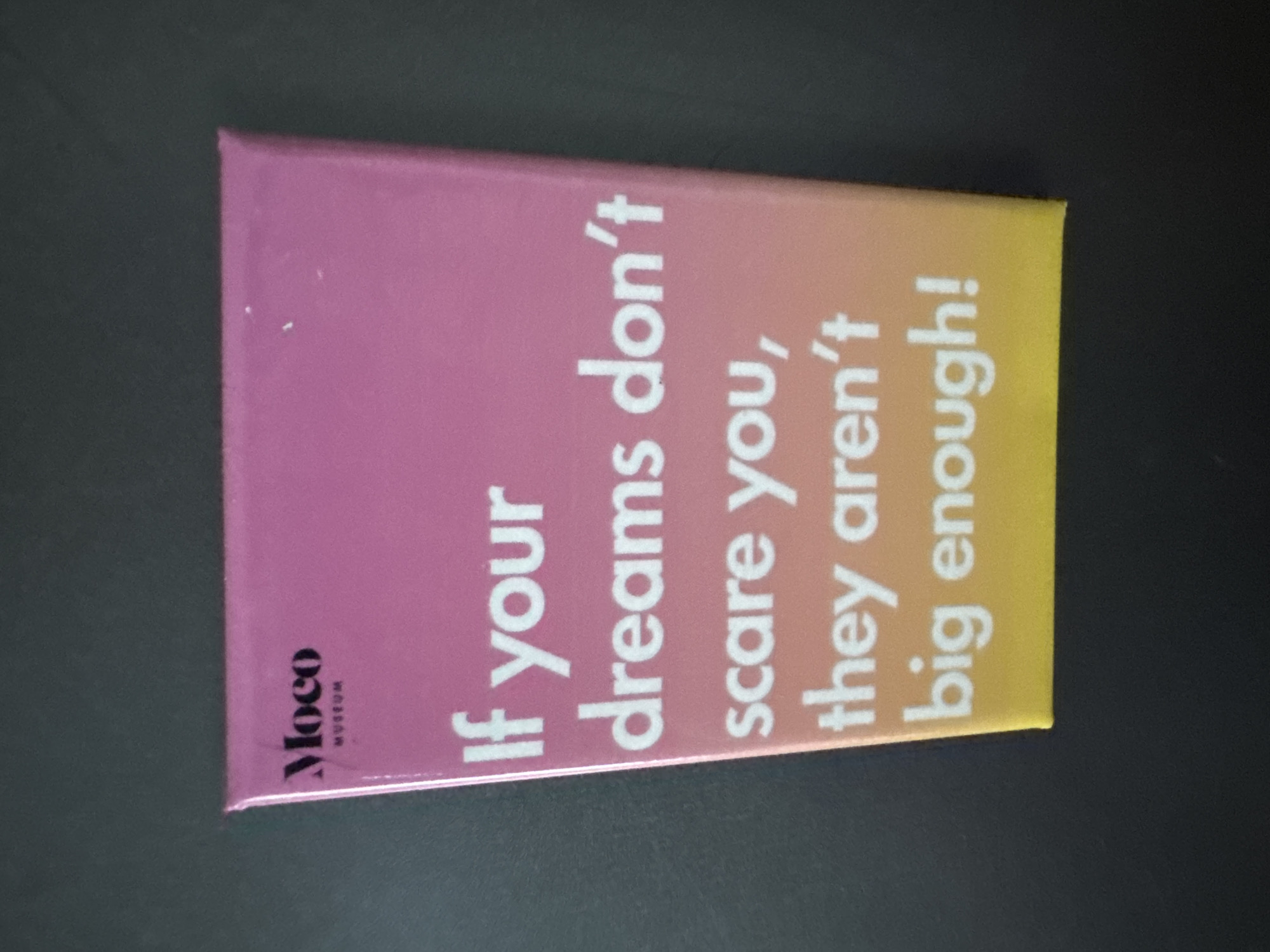 The image shows a postcard from the Moco Museum. The postcard is divided into three color sections: the left side is pink, the middle section is a gradient of orange, and the right side is yellow. The text on the postcard reads, "If your dreams don't scare you, they aren't big enough!" The Moco Museum logo is in the lower left corner. The postcard is placed on a dark surface.
