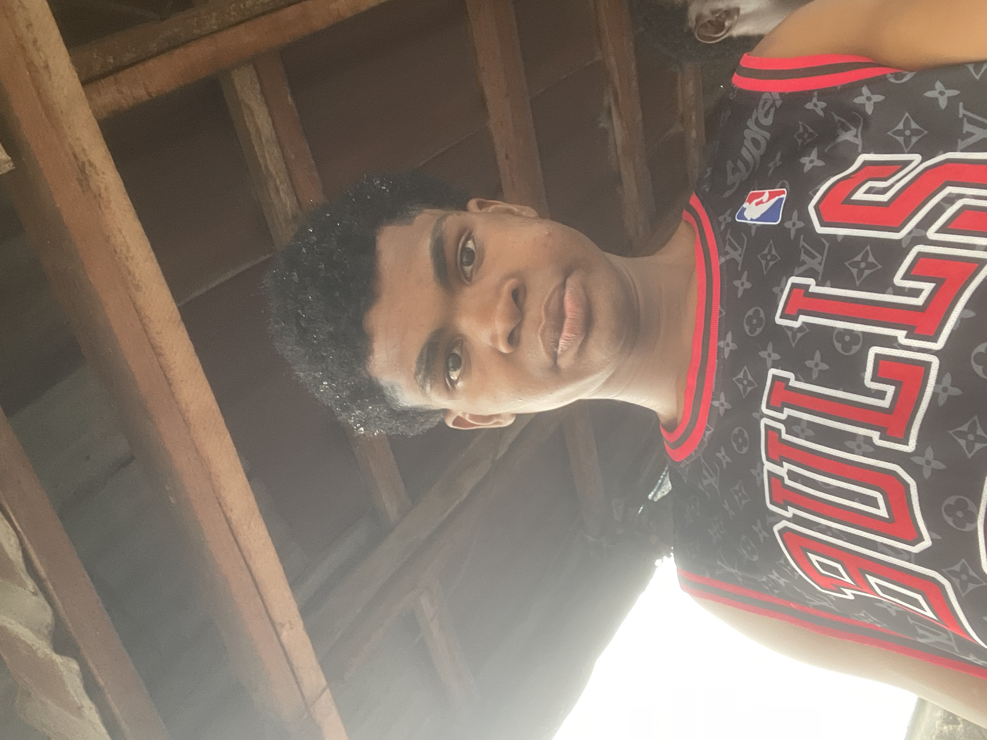 A young Black male is pictured from the chest up, looking directly at the camera. He has dark, curly hair and dark eyes. He is wearing a black basketball jersey with "BULLS" written in red and white letters across the chest. The jersey also has a repeating pattern of a Louis Vuitton-like monogram and the NBA logo. The background shows wooden beams of a roof structure, suggesting an outdoor or covered area. The lighting is bright, with a strong light source coming from the left, creating a hazy effect. The image appears to have been taken in Ado, Nigeria.
