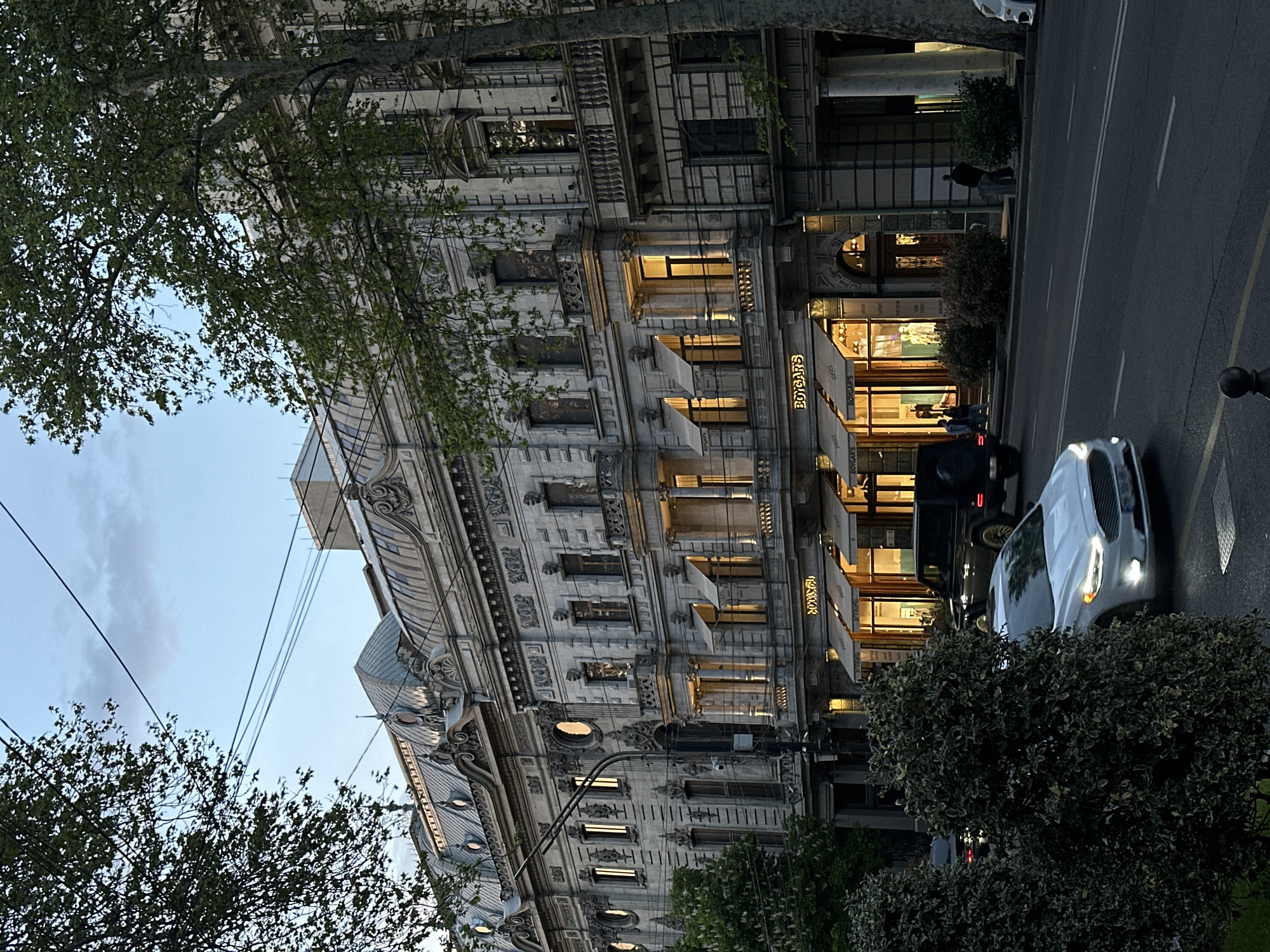 A street-level view, taken near dusk in Tbilisi, Georgia, features a Beaux-Arts style building that houses the boutique "BOYARS". The building's facade is decorated with intricate architectural details, including pilasters, cornices, and ornamental carvings. Windows are illuminated with warm light, reflecting on the awnings bearing the "BOYARS" name. A street runs parallel to the building, featuring vehicular traffic consisting of a white car and a black SUV. Pedestrians are walking near the entrance of the store. Foliage from trees and bushes frames the shot on the left and right sides of the frame. Overhead electrical cables stretch across the sky.