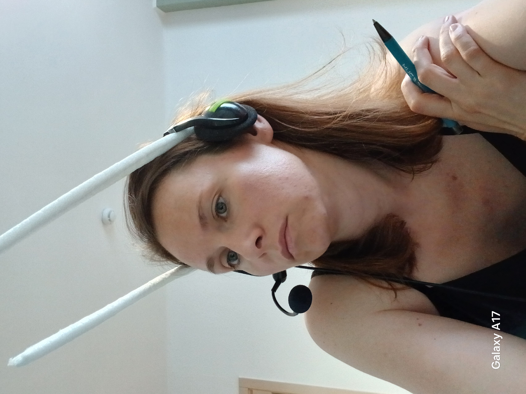 A woman with reddish-brown hair is looking at the camera. She is wearing a headset with a microphone. She is holding a blue pen in her right hand. Two white, cylindrical objects are above her head. The picture was taken by a Galaxy A17 smartphone. The location is unknown.