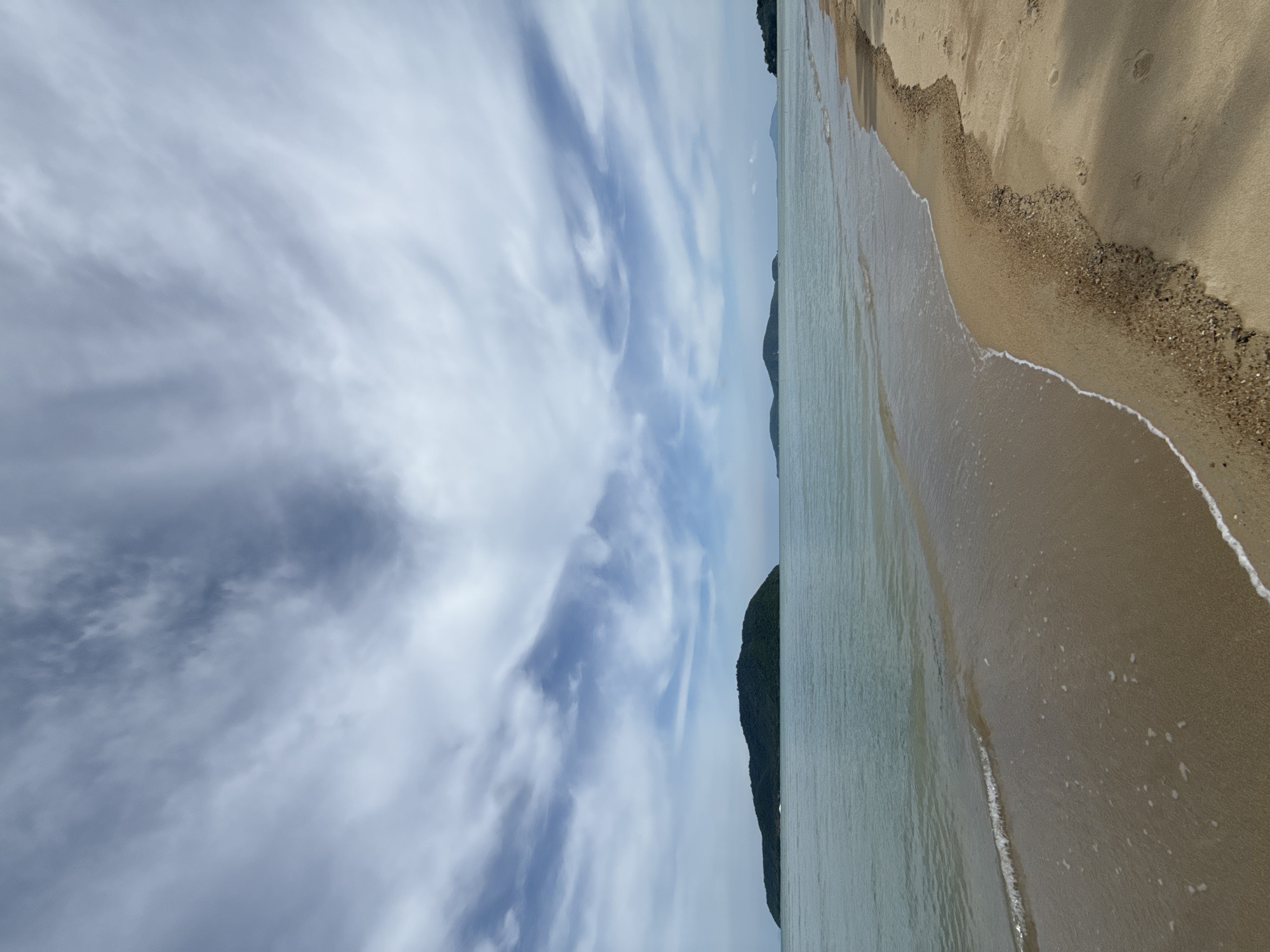 The image depicts a wide, sandy beach with gentle waves lapping at the shore. In the distance, several lush, green islands are visible, silhouetted against a sky filled with dramatic, wispy clouds. The water is a tranquil blue-green, and the sand is a light brown, showing a few footprints. The scene appears to be a serene coastal landscape. This is a view of the beach in Rawai, Thailand.