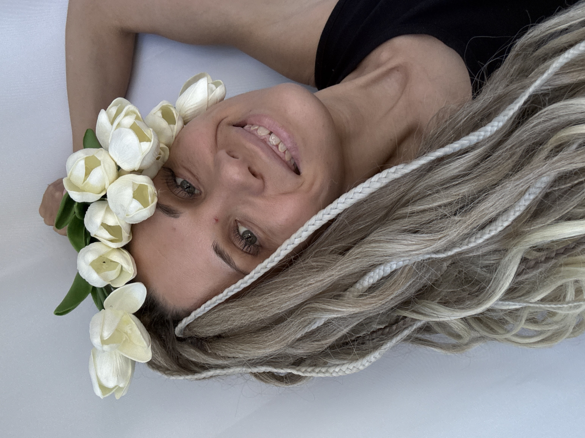 A woman with light skin and blonde hair with white braids lies on a white surface. She is wearing a black shirt and a flower crown of white tulips on her head. She is smiling and looking directly at the camera. The location is not discernible from the image.