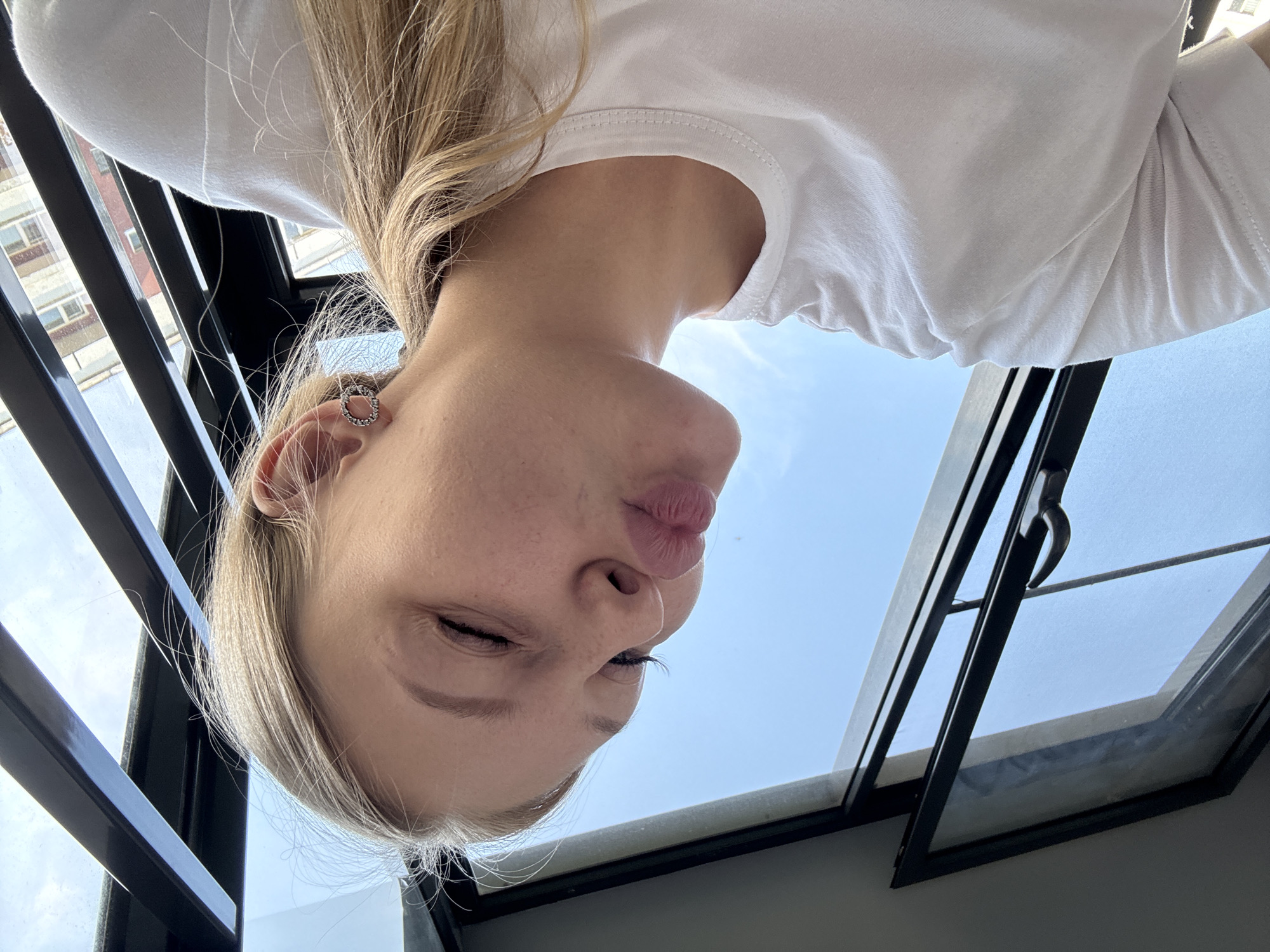 A young woman with blonde hair pulled back into a ponytail is captured in a selfie, puckering her lips for a kiss. She is wearing a plain white t-shirt and a circular earring with small rhinestones in her left ear. She is positioned in front of a large window with dark frames, through which a bright blue sky with some wispy clouds is visible. The window appears to be part of an apartment building or office space. The background shows a glimpse of other buildings. The scene is brightly lit by natural light.