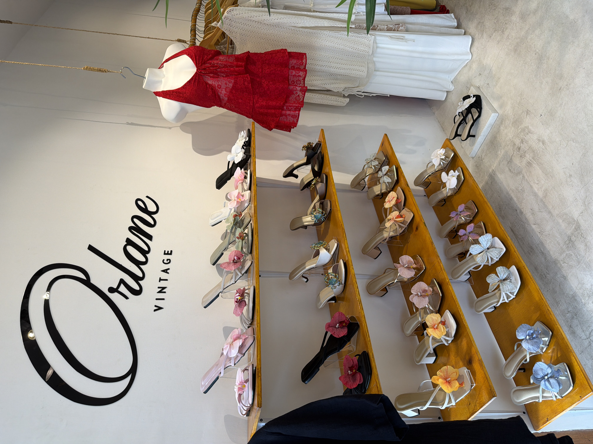 A retail store is captured in a slightly tilted full shot, possibly in Kuta, Indonesia, based on the presence of the name "Orlane Vintage," which may be a local business. The interior wall is adorned with the shop's logo on a white background. A rack of shoes displayed on angled wooden shelves dominates the scene. The footwear is primarily sandals in varying colors, some decorated with floral ornaments. Garments, including a red dress and white attire, are hung higher in the frame. The flooring appears to be concrete. The overall impression is that of a well-organized boutique setting.
