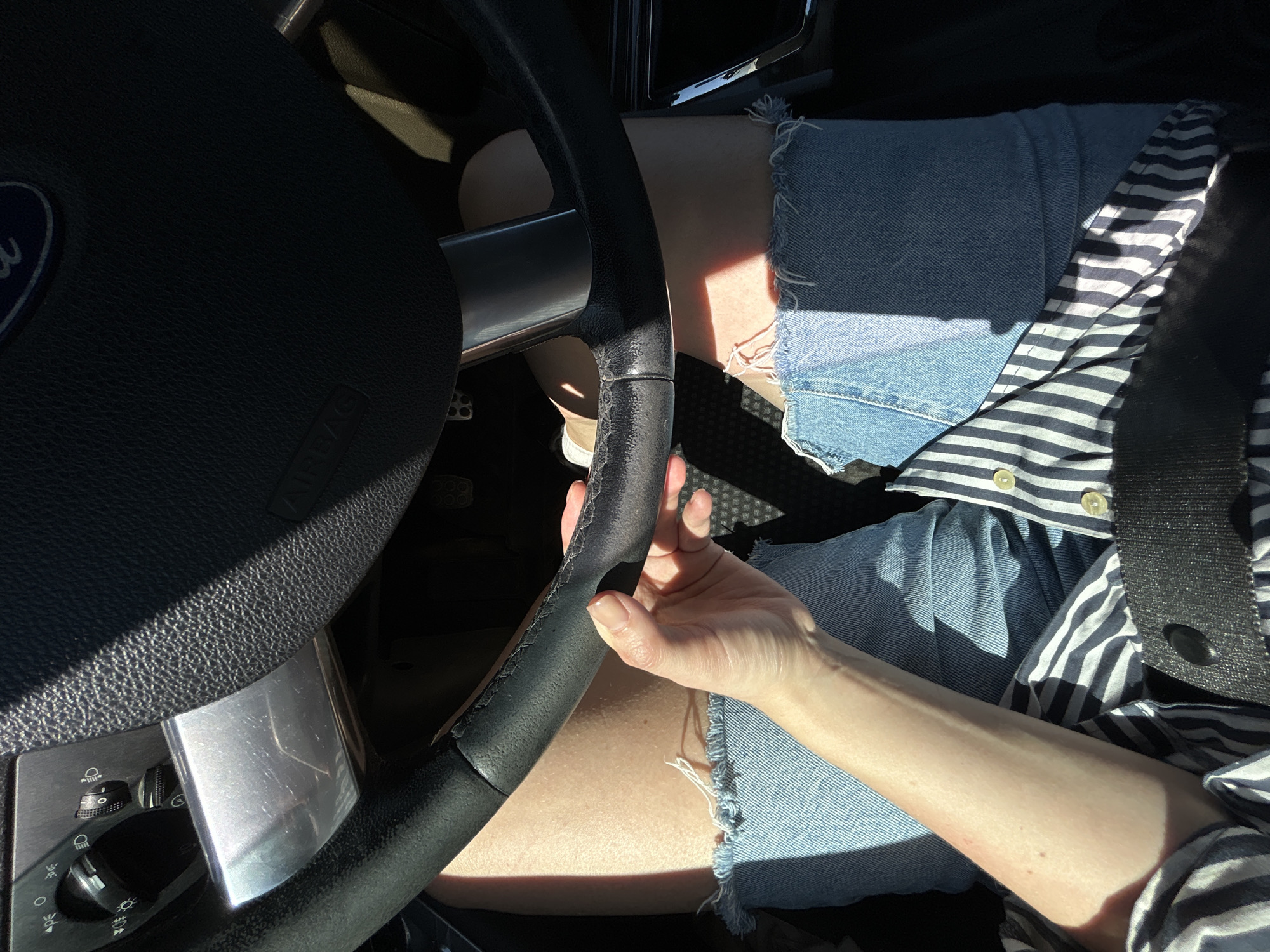 The image shows a close-up view of a person's hands and legs inside a car, as seen from the driver's perspective. The driver, likely female based on the hands and legs, is wearing denim shorts and a striped shirt. The hands are gripping the steering wheel, which appears to be black leather. The car interior is visible, including part of the dashboard with light controls and a silver accent piece. Sunlight streams in, casting strong shadows across the scene. The setting is not specified as being in the city of Ленинское, Russia.