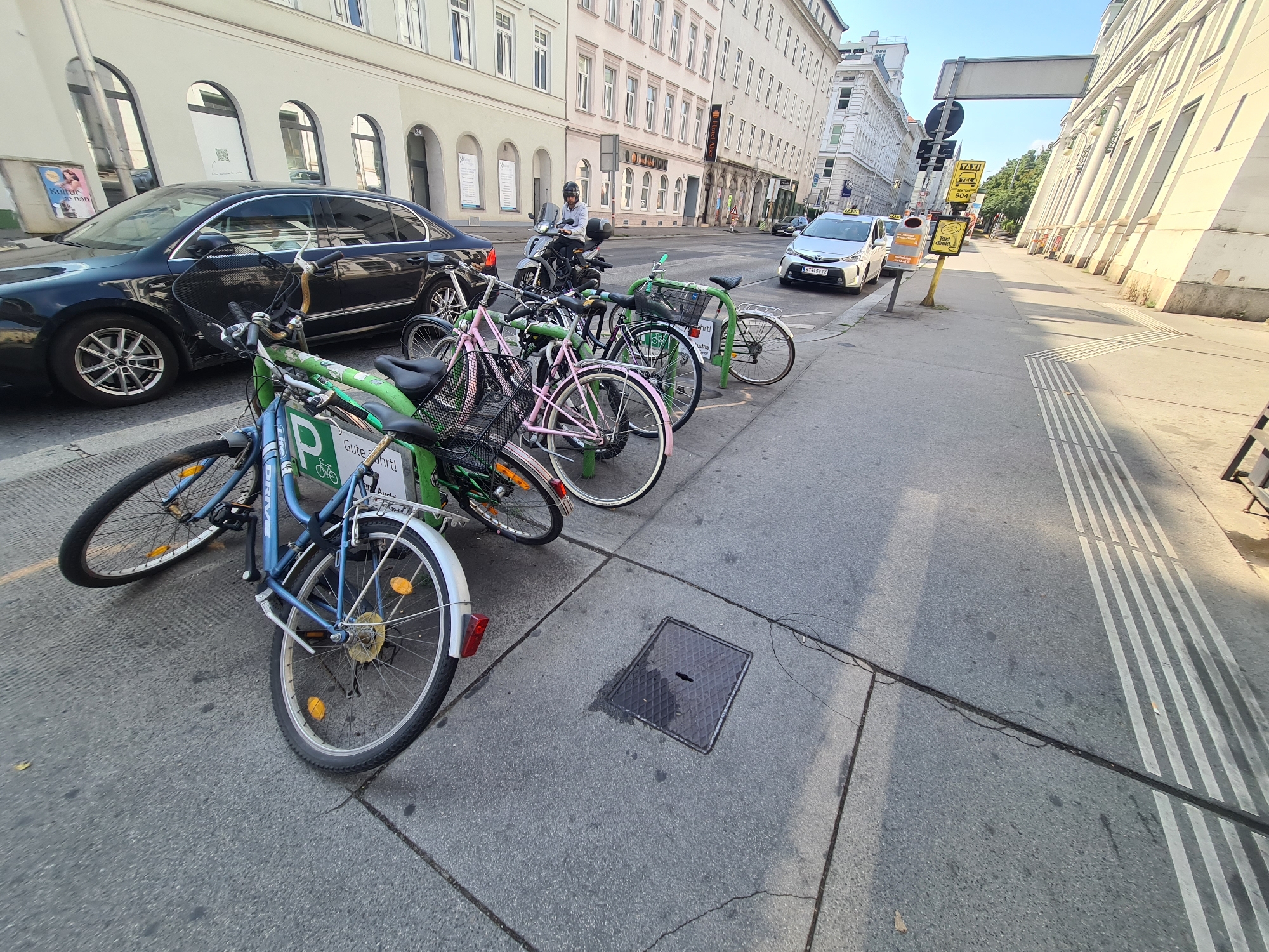 This is a ground-level, eye-level, medium shot of a row of parked bicycles on a city sidewalk in Vienna, Austria. The bicycles are of various colors, including blue, green, and pink, and appear to be part of a rental service, as indicated by the prominent "P" logo on one of the blue bikes and a green rack with a sign that says "Gute Fahrt!" (Have a good trip!). The bicycles are arranged in a line along a street, with a dark-colored sedan parked to the left and a white taxi driving by in the background. The street is paved with asphalt, and the sidewalk has a textured surface with raised white stripes, suggesting it is a pedestrian crossing or tactile paving for the visually impaired. The buildings in the background are typical of Viennese architecture, with tall, multi-story structures featuring ornate windows and balconies. The sky is clear and blue, indicating a sunny day. There is also a motorcycle with a rider dressed in a helmet and light-colored jacket, parked near the bicycles. The overall scene depicts a typical urban streetscape in Vienna, with elements of transportation and daily life.