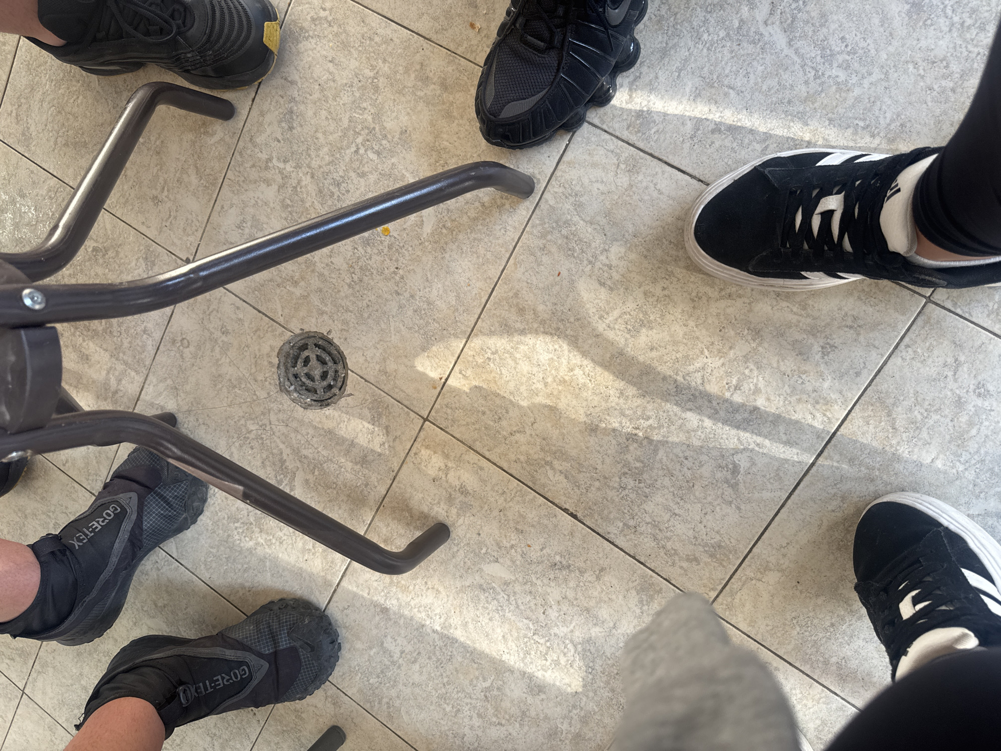 The image shows a tiled floor with four pairs of shoes and the legs of a chair. The shoes are of various styles and colors: one pair is black with a yellow sole, another is entirely black, a third is black with white stripes, and the fourth is black with "GORE-TEX" written on the side. The chair legs are made of a dark metal. The location is unknown.