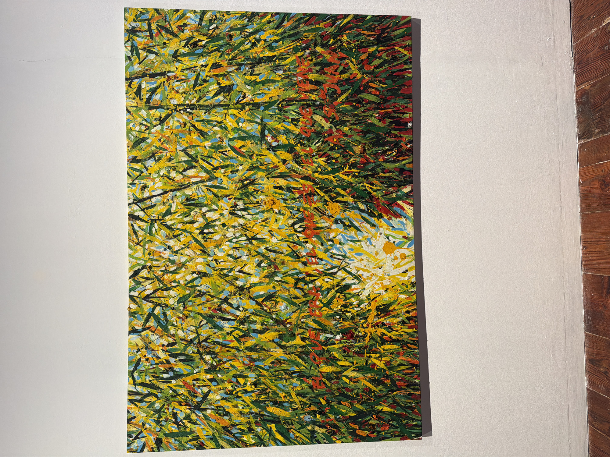 This is a vertical, close-up, eye-level shot of a vibrant abstract painting hanging on a textured, off-white wall. The painting is a dense tapestry of short, expressive brushstrokes in various shades of green, yellow, orange, red, and blue. The strokes are layered and overlapping, creating a sense of movement and depth. There are hints of black and white interspersed, adding further visual complexity. The overall impression is one of a chaotic yet harmonious natural scene, perhaps suggesting foliage in a strong wind or a sun-drenched field.
The painting occupies the right half of the frame. The left side of the image is dominated by the plain, off-white wall, which exhibits subtle texture and a few faint imperfections. To the far right of the painting, a narrow section of a wooden wall is visible, with dark, vertical planks showing a natural wood grain and some visible knots. The lighting appears to be artificial, casting a soft glow on the artwork and the wall. There is no visible text or any depiction of people or specific objects. The setting suggests an indoor display, possibly an art gallery or studio space, given the presentation of the artwork and the neutral background.
