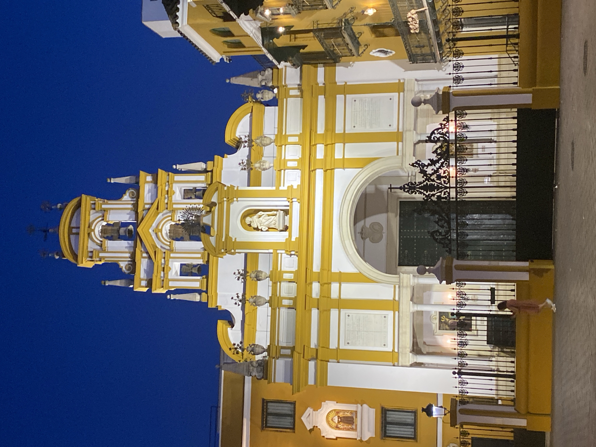 The image depicts the Iglesia de la Magdalena in Seville, Spain, at dusk. The church's facade is painted in bright yellow and white, with ornate architectural details. A central arched entrance is flanked by columns and decorative elements. Above the entrance, a niche contains a statue of a saint. The church tower, visible in the upper part of the image, is also elaborately decorated with yellow and white accents and topped with crosses.
To the left of the church, a yellow building with windows is visible, and a small niche with a statue of a nun is embedded in its facade. To the right, another yellow building with balconies and arched windows is visible.
In the foreground, a black wrought-iron fence separates the church from the street. A person wearing a light-colored dress is walking along the sidewalk in front of the fence. The street is paved with cobblestones. The sky above is a deep blue, indicating the late hour. The scene is illuminated by streetlights and interior lighting from the church and surrounding buildings.