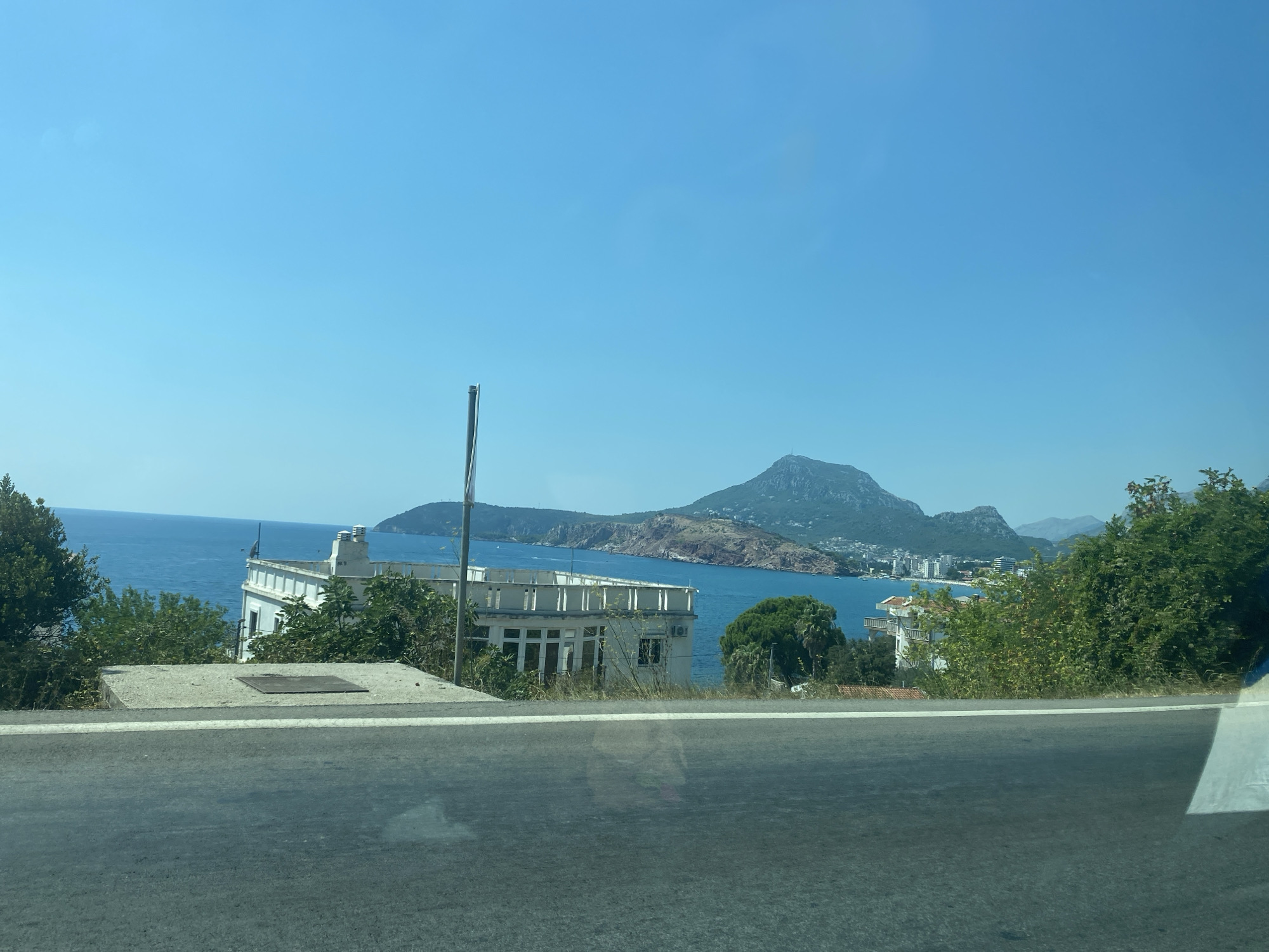 This is a photograph taken from a vehicle, showing a coastal landscape under a clear blue sky. In the foreground, a grey asphalt road with a white line marking its center runs horizontally across the frame. To the left of the road, a rectangular concrete structure with a metal plate embedded in its surface is visible.
Beyond the road and concrete structure, a white building with a flat roof and a railing on its balcony is situated on a slight incline. This building overlooks the sea, which is a vibrant blue. Lush green trees and vegetation surround the building and extend down towards the coastline.
In the middle ground, the coastline curves, revealing more buildings and vegetation on the hillsides. A prominent, large mountain dominates the background, with a smaller, rounded mountain to its left. The distant mountains appear to be part of the Sutomore, Montenegro region, as suggested by the context. The overall scene depicts a sunny day in a Mediterranean coastal town.