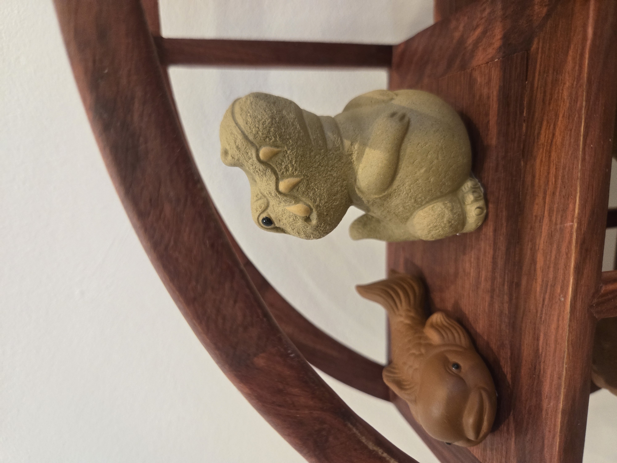 A close-up shot shows two clay figures attached to a wooden structure. The wooden structure is dark reddish-brown and appears to be part of a shelf or decorative piece with curved elements. The clay figures include a light-brown hippopotamus-like creature on top, with small protruding horns and black eyes. Below it, a similarly-colored fish-shaped figure is attached to the wood. The background is a plain white wall. There is no evidence in the picture to suggest this image was taken in Budva, Montenegro.