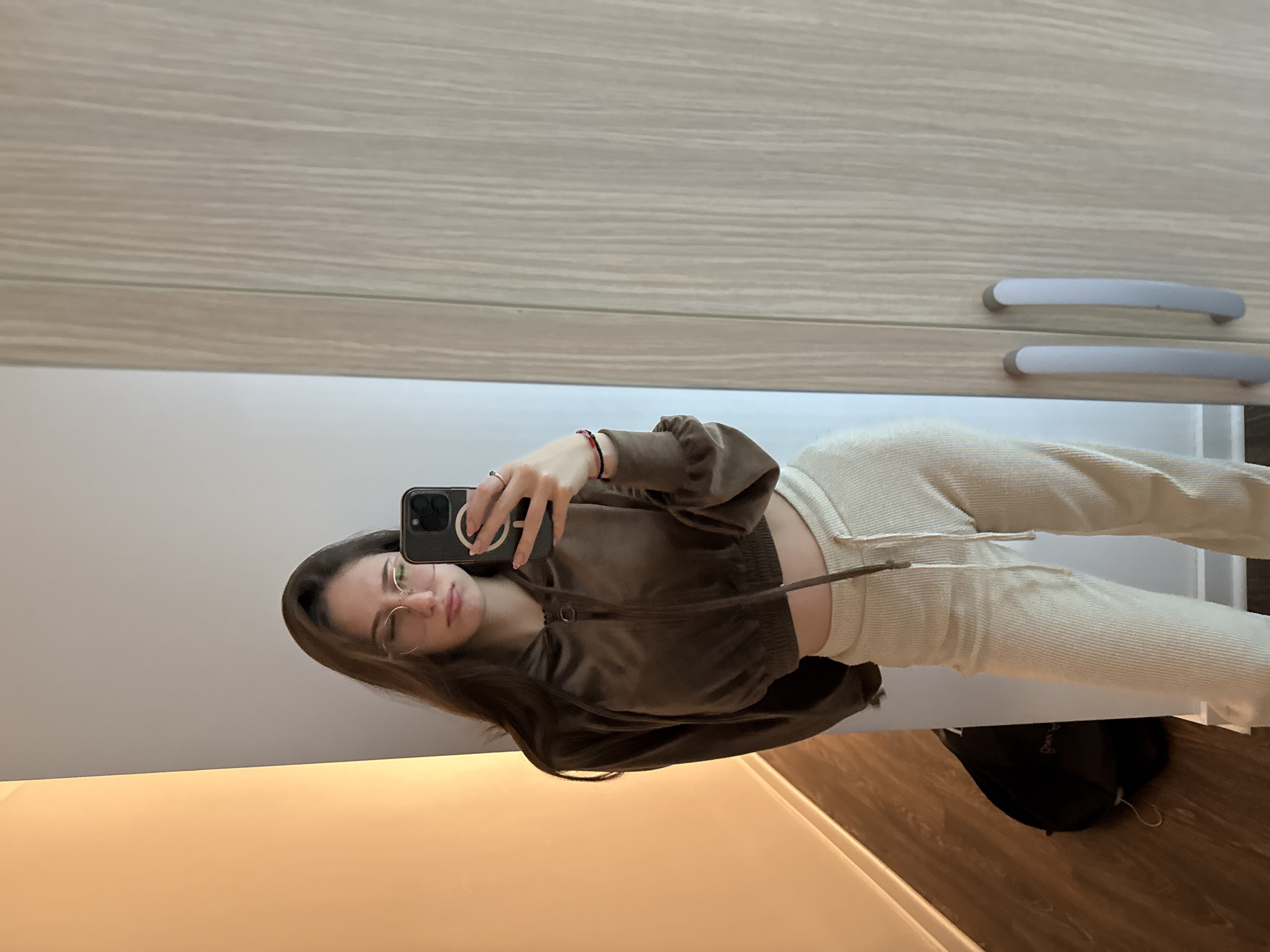 A female individual is depicted taking a mirror selfie in a domestic interior located in the Vozdovac Urban Municipality, Serbia. She has long, dark brown hair, wears round, light-rimmed glasses, and exhibits a slight pout with furrowed eyebrows. She is dressed in a dark brown, velvet-like cropped hoodie with a zipper and drawstrings, paired with light beige ribbed lounge pants secured with a drawstring. A ring is present on her left ring finger, and a red and black bracelet on her left wrist. She holds a dark-colored smartphone, featuring a pronounced camera bump and a visible magnetic ring on its case, with her left hand, aiming it towards the mirror. The background, largely a mirror reflection, shows a plain light grey wall behind the subject. To the left, a vertical section of wall exhibits a warm, light beige-orange coloration, below which a dark brown wooden floor is visible. A dark backpack rests on the floor against the wall. The immediate right side of the image displays a light wood-grain wardrobe or closet with two silver-colored, curved handles. The scene is illuminated by indoor lighting.