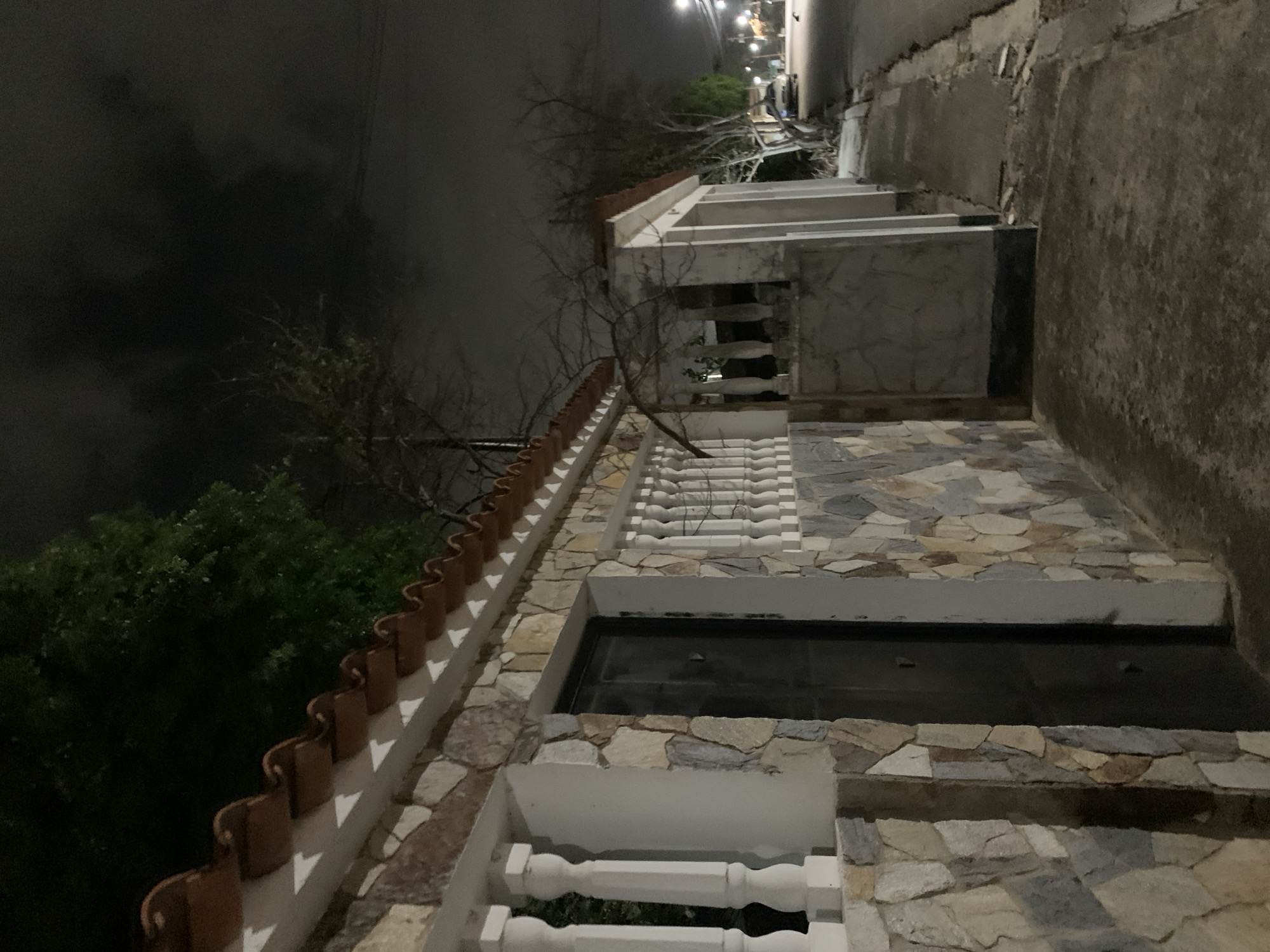 The image captures a steep, multi-level outdoor pathway or staircase ascending a hillside at night in Santa Ana, Venezuela. On the right, a series of structures or buildings lines the incline, featuring walls clad with irregular stone tiles in various shades of grey, beige, and brown. White, decorative balustrades serve as railings or balcony fronts on different levels. Some sections include dark, rectangular recesses that appear to be windows or sheltered areas. Along one upper edge, terracotta-colored, half-round roof tiles are used as a decorative coping. The path itself seems to be paved with similar stone tiles.
To the left of the structures, the sky is dark and heavily clouded, with tree branches, some bare and some with green foliage, silhouetted against it. In the distance, at the top of the incline, artificial lights glow, indicating further development or a street. The overall lighting is dim, characteristic of nighttime, with light sources mainly coming from the distance and perhaps subtle illumination on the structures themselves. There are no people visible, and no specific activity is taking place. The scene evokes a quiet, residential atmosphere in a hilly terrain. No visible text is present in the image.