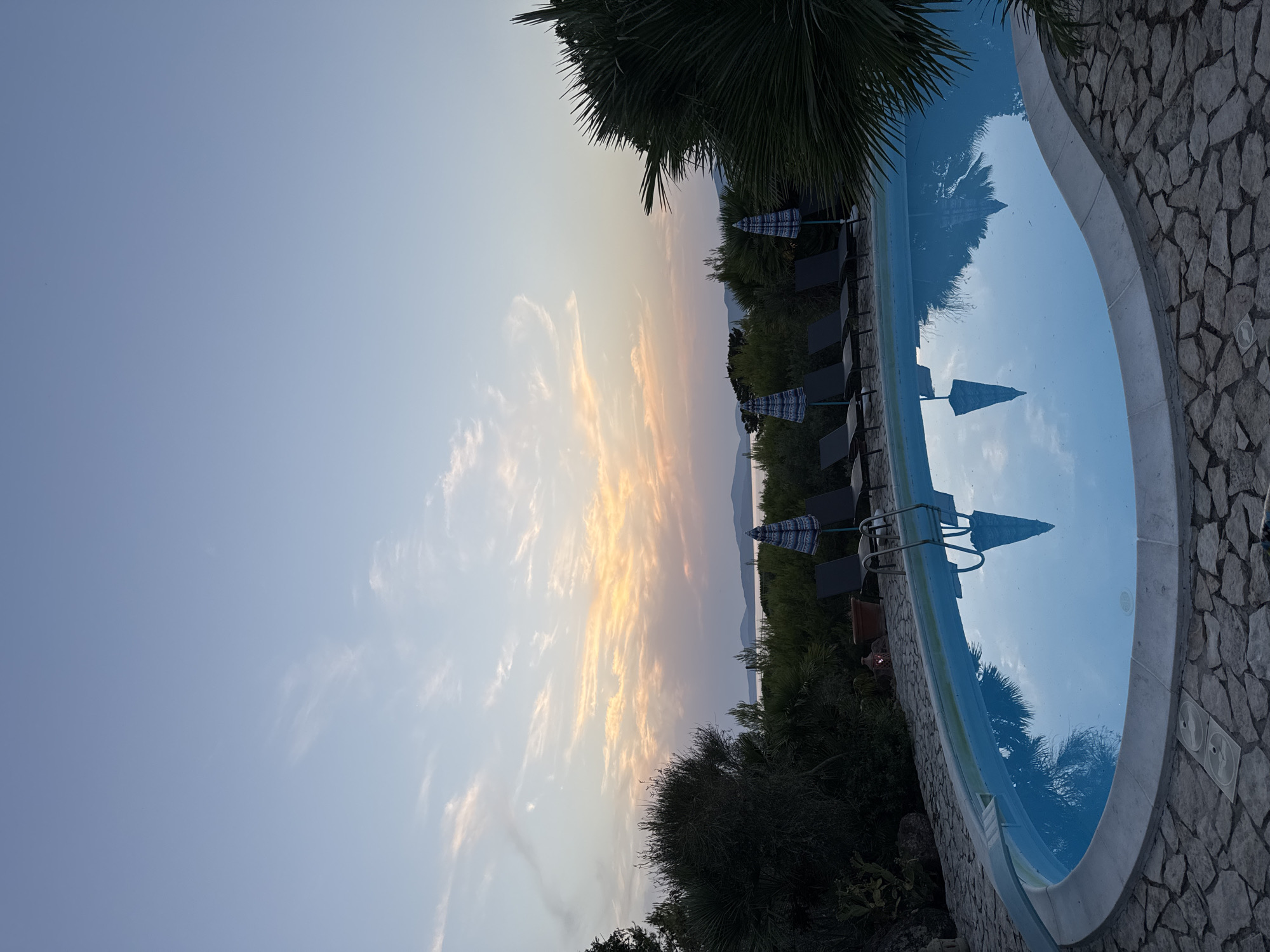 The image depicts an outdoor swimming pool at dusk, situated in a setting that suggests a coastal location. The pool is kidney-shaped and filled with clear blue water, reflecting the sky and surrounding elements. The pool is bordered by a stone patio.
To the right of the pool, lounge chairs and umbrellas are visible. Behind the pool and lounge chairs, there is dense vegetation, including trees and palm fronds, with a body of water and a distant coastline visible on the horizon under a sky painted with the soft colors of sunset. The overall impression is one of a tranquil, serene evening scene.
While the image showcases a beautiful setting, it does not contain enough specific visual cues to definitively pinpoint its exact location within Alghero, Italy, or to identify any specific individuals or activities.
