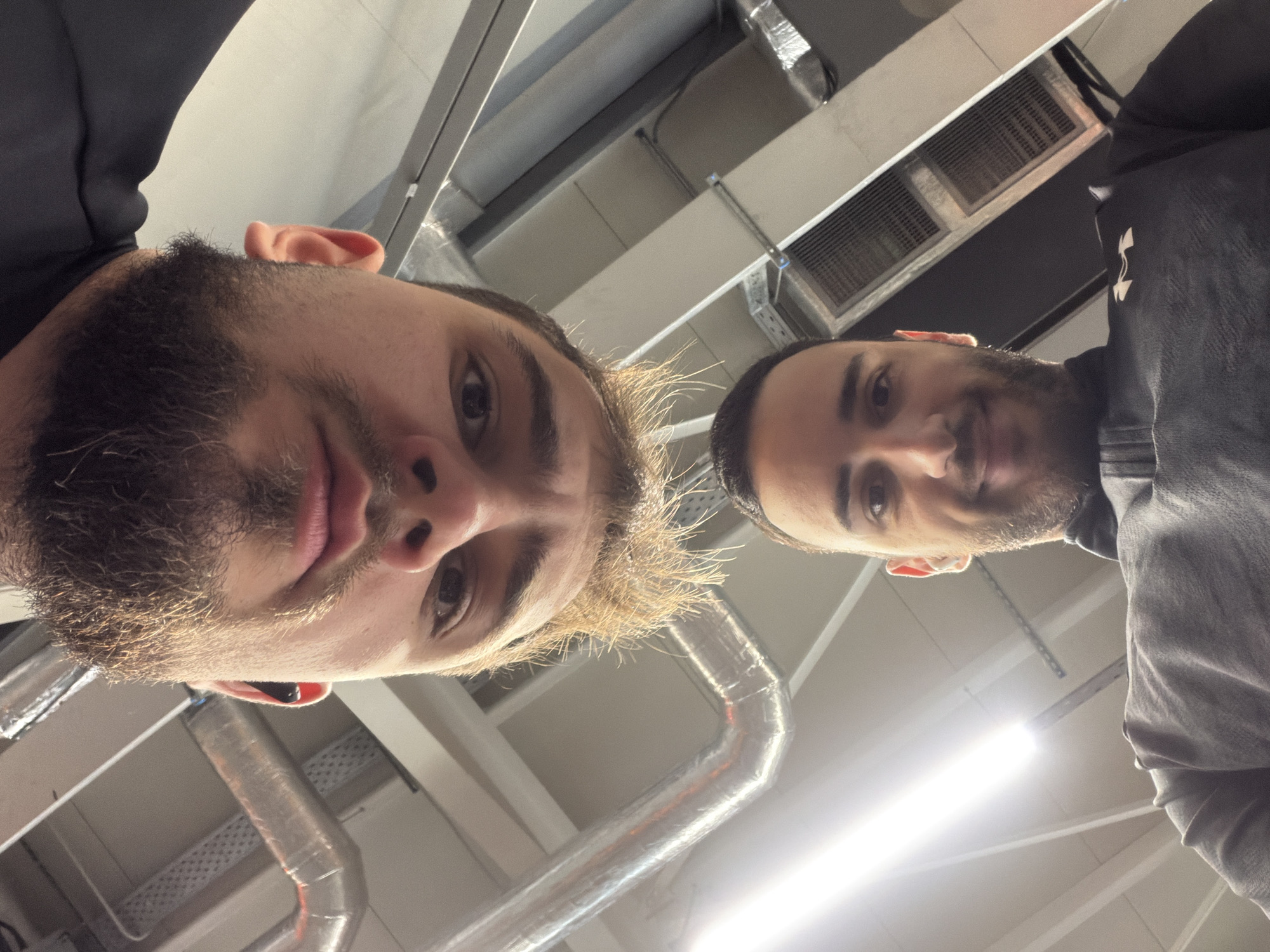 Two men are posing for a selfie in an industrial-looking space. The man on the left has fair skin, a full beard, and light brown hair. He is wearing an earbud. The man on the right has darker hair and a trimmed beard. The location appears to be indoors, with exposed pipes and ventilation systems in the ceiling. It is not possible to pinpoint the location within Plovdiv, Bulgaria, with the available information.