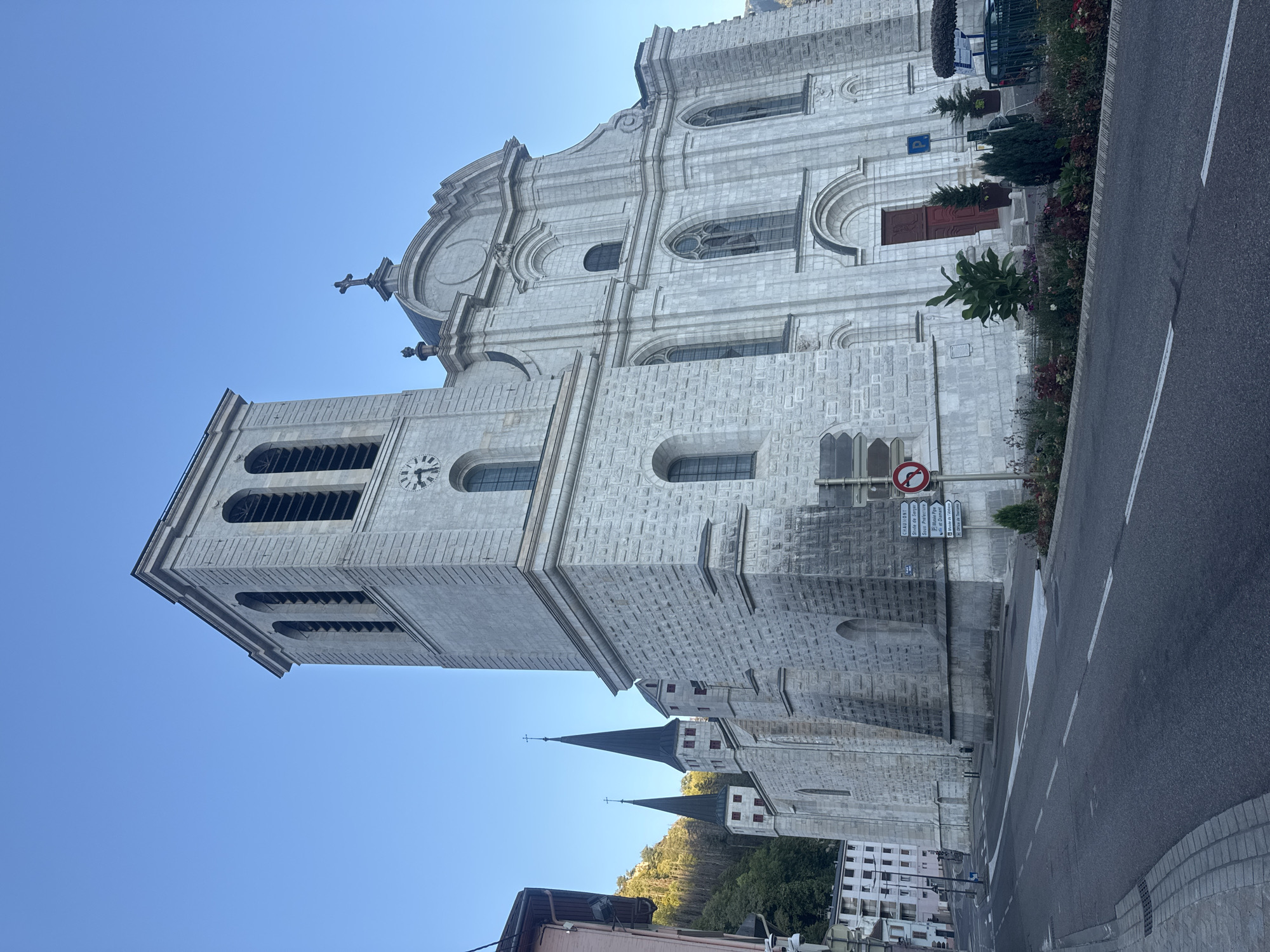 The image depicts a large, stone church in the city of Saint-Claude, France. The church has a prominent bell tower with arched openings and a clock face. The facade of the church is ornate, with multiple arched windows and a large red double door. Two tall, dark spires with crosses on top rise from the rear of the church. The church is situated on a street with a sidewalk and road. In the background, a wooded hill and several buildings are visible. There are no people visible in the image. The scene is bathed in sunlight under a clear blue sky.