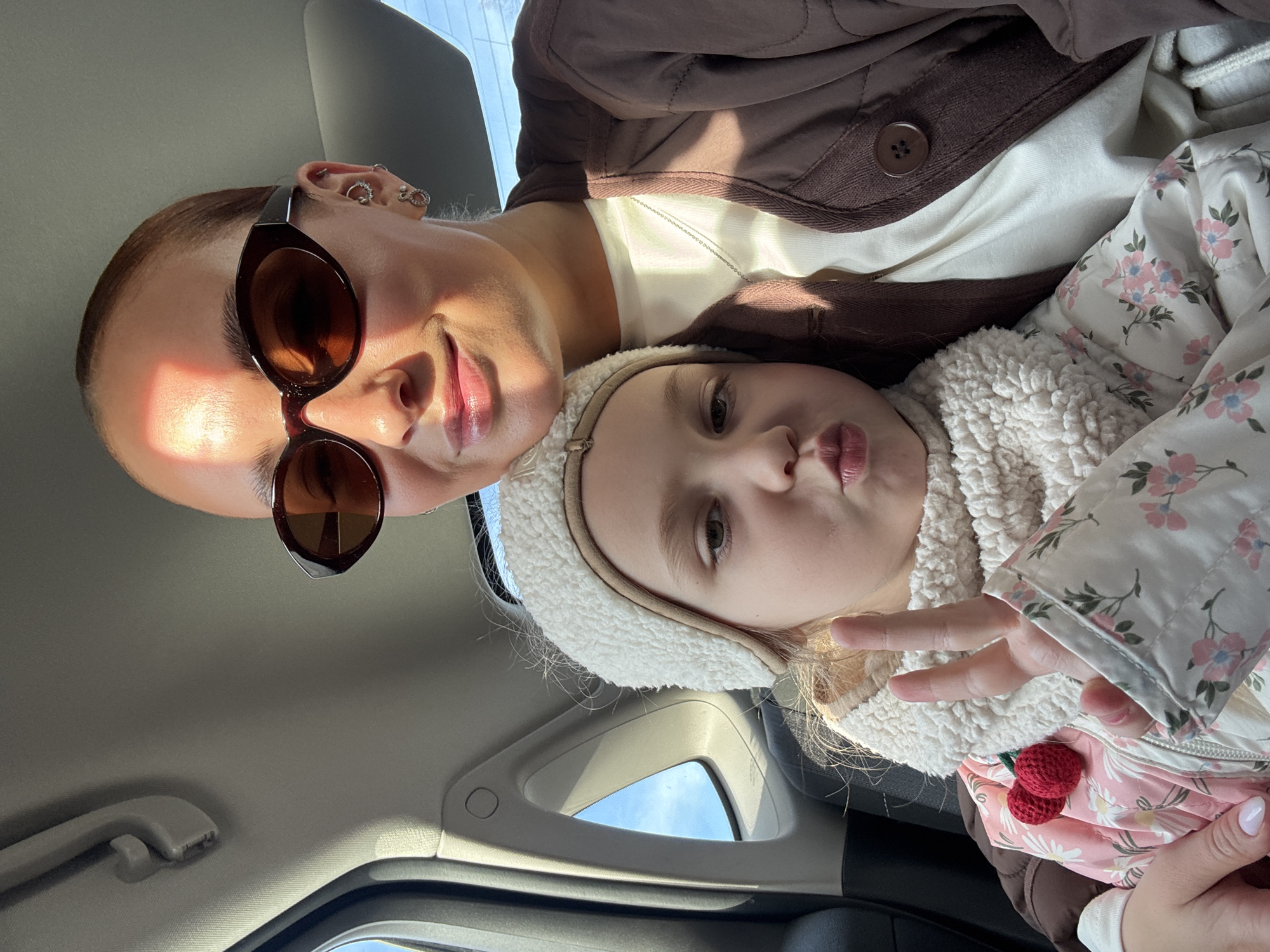 A woman with short hair, brown sunglasses, and multiple ear piercings is taking a selfie with a child. The child is wearing a white, fur-lined hat and a floral-patterned coat with a crochet cherry attached. The child is making a peace sign. The photo appears to be taken inside a car. The location, Chaikovsky, Russia, cannot be verified from the image.
