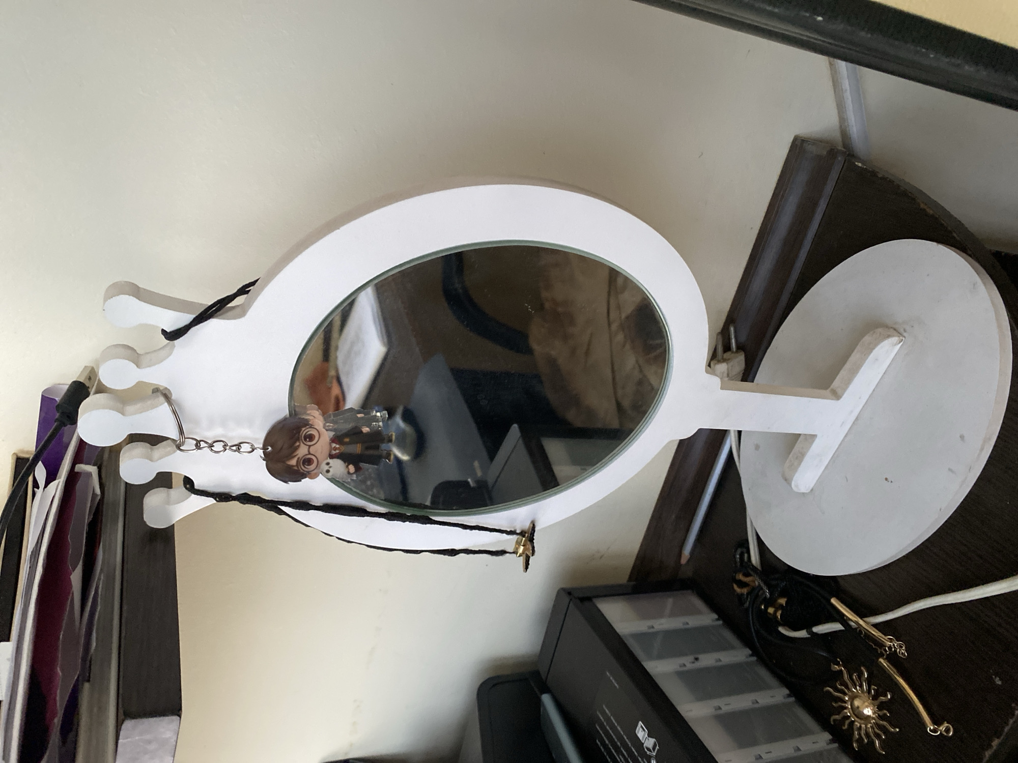 A white decorative mirror, shaped like a crown, stands on a flat white base. A black cord hangs from one of the crown points, with a small keychain featuring a cartoon character resembling Harry Potter holding an owl attached to it. Below the mirror, on a dark surface, lies a gold sun-shaped pendant with radiating rays, attached to a black cord and gold chain. To the left of the mirror is a black electronic device with compartments. The background consists of a plain, light-colored wall and a shelf above, holding various papers and dark objects. The scene is indoors, with artificial lighting.