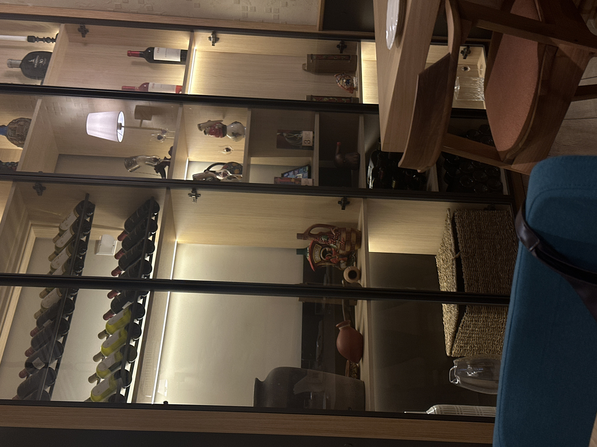 The image shows an interior scene, possibly a restaurant or a private dining area, featuring a display cabinet filled with various items. The cabinet is illuminated from within, highlighting the contents on its shelves.
On the top shelf of the left section of the cabinet, there are several rows of wine bottles, suggesting a wine cellar or collection. Below this, the shelves contain decorative items, including a large dark vase on one shelf, a red ceramic object that appears to be a jug or bottle, and other smaller decorative pieces.
The right section of the cabinet also displays wine bottles on the top shelves. Further down, there are decorative items such as a small lamp with a white shade, glass decanters, and figurines. The lower shelves hold more bottles and decorative items, some of which appear to be antique or collectible.
In the foreground, a wooden table and chair are visible, indicating a dining or seating area. A white plate rests on the table. The overall ambiance is dimly lit, with warm lighting emanating from the display cabinet and possibly from other sources not fully visible in the frame.
There are no people visible in the image. The environment appears to be indoors, with a wall featuring a textured pattern visible in the background. The materials of the display cabinet appear to be wood and glass.
The image is consistent with an indoor setting in Belgrade, Serbia, likely a restaurant, bar, or a home with a well-appointed wine collection and display.