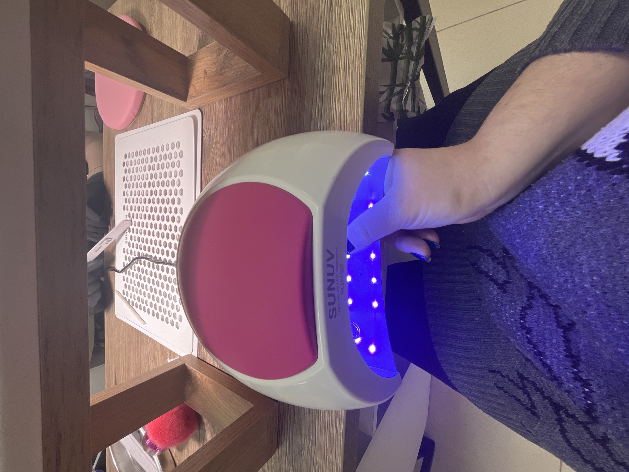 The image depicts a SUNUV brand UV LED nail lamp in use, with a hand placed inside to cure nail polish. The lamp is white with a pink interior and is emitting a blue light from the UV LEDs. The hand has manicured nails with blue nail polish. The lamp is positioned on a wooden surface, possibly a table or desk. To the left of the lamp is a white object with a grid pattern. There is a pink object visible beneath the table. The person using the lamp is wearing a dark gray garment with a geometric pattern. The location of the scene is unknown and there is no evidence suggesting it is in Klin, Russia.