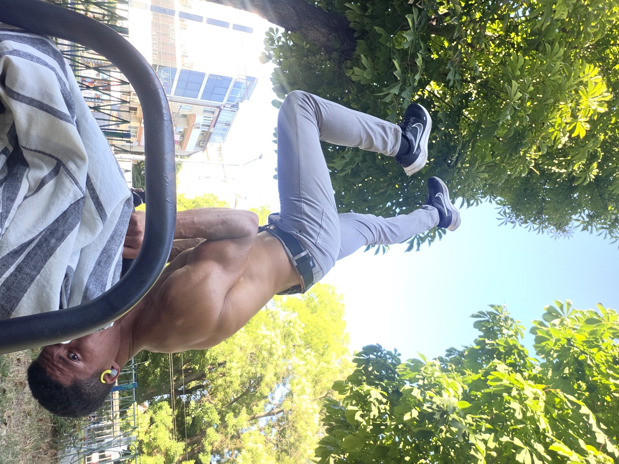 A shirtless man with dark skin is performing a handstand outdoors, supported by his hands on a metal bar. His body is positioned upside down, with his legs extended upwards and his feet wearing black and white athletic shoes. He is wearing light-colored pants with a dark belt. His head is tilted downwards, and he is looking towards the camera. He has a yellow earbud in his left ear. The background shows lush green trees with dappled sunlight, a clear blue sky, and a building with blue-tinted windows in the distance. A striped cloth is draped over the metal bar in the foreground. The scene appears to be in an urban park setting, potentially near a playground given the metal bar and the presence of grass and trees. This specific location is in the city of Varna, Bulgaria, near the Primorski Park.