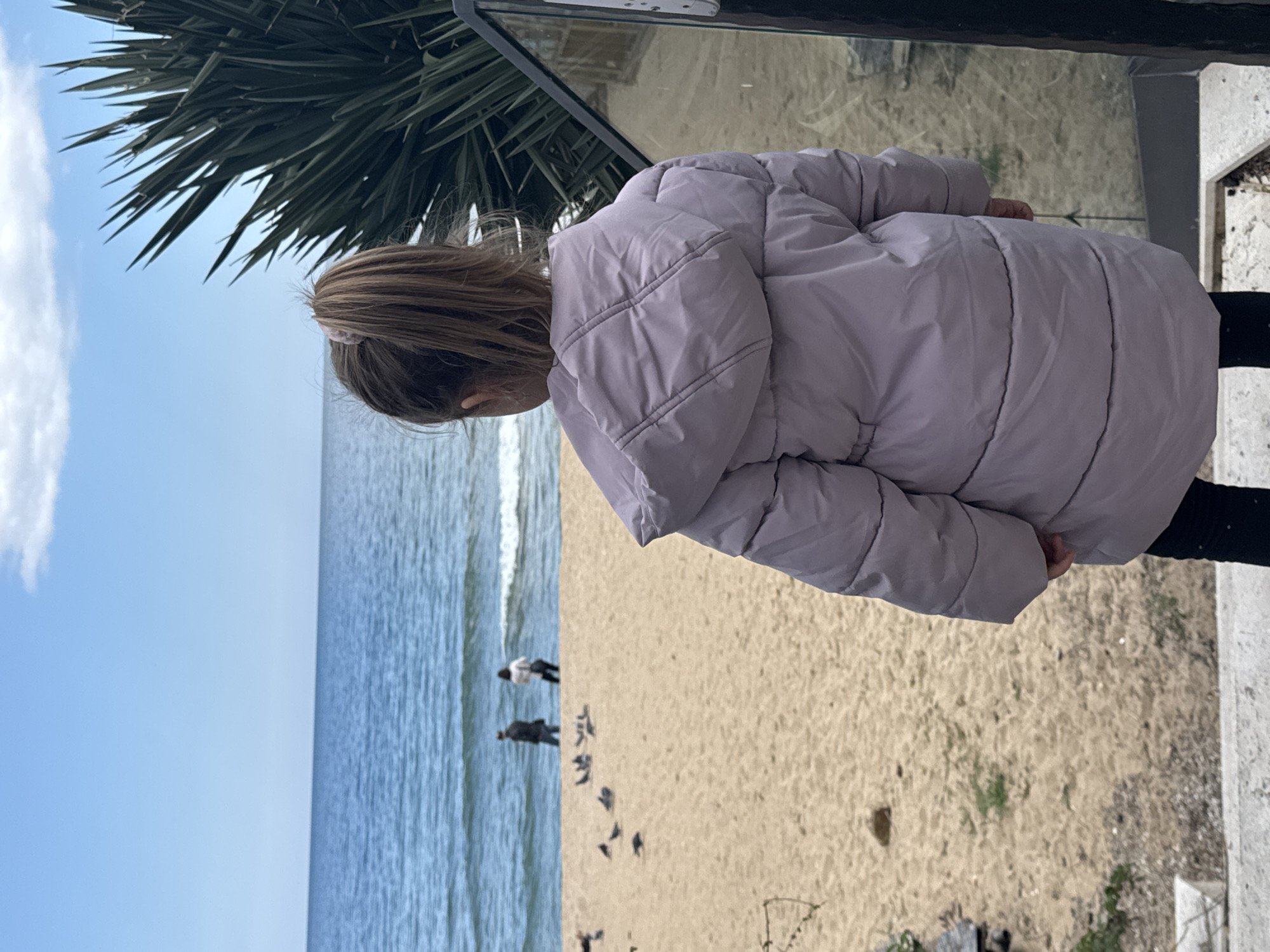 A young girl with brown hair tied back stands on a concrete structure overlooking a beach in Varna, Bulgaria. She wears a puffy lavender jacket and black pants, looking down towards the sandy beach. The Black Sea stretches to the horizon under a blue sky with scattered clouds. Two people walk along the water's edge. Several birds stand and peck in the sand. A palm tree extends into the frame from the upper left. A glass barrier separates the girl from the scene below.