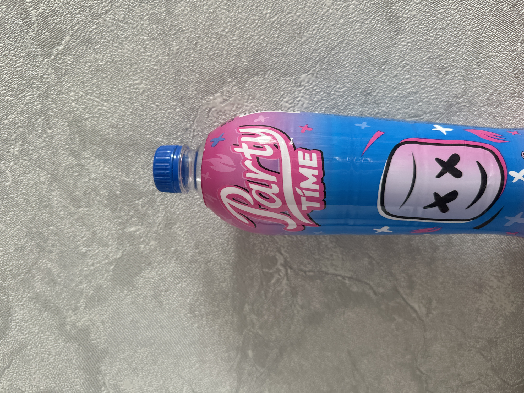 A close-up, eye-level shot shows the top portion of a blue and pink plastic bottle with a blue cap. The bottle is labeled "Party Time" in stylized white and pink lettering, with abstract pink and blue graphics that resemble flames and stars. A graphic of a white marshmallow with black "X" eyes and a smiley mouth is also visible on the bottle. The bottle is placed against a textured, light-colored wall. The scene appears to be indoors, possibly in a room or on a shelf, and there is no indication of any specific location in Birobidzhan, Russia.