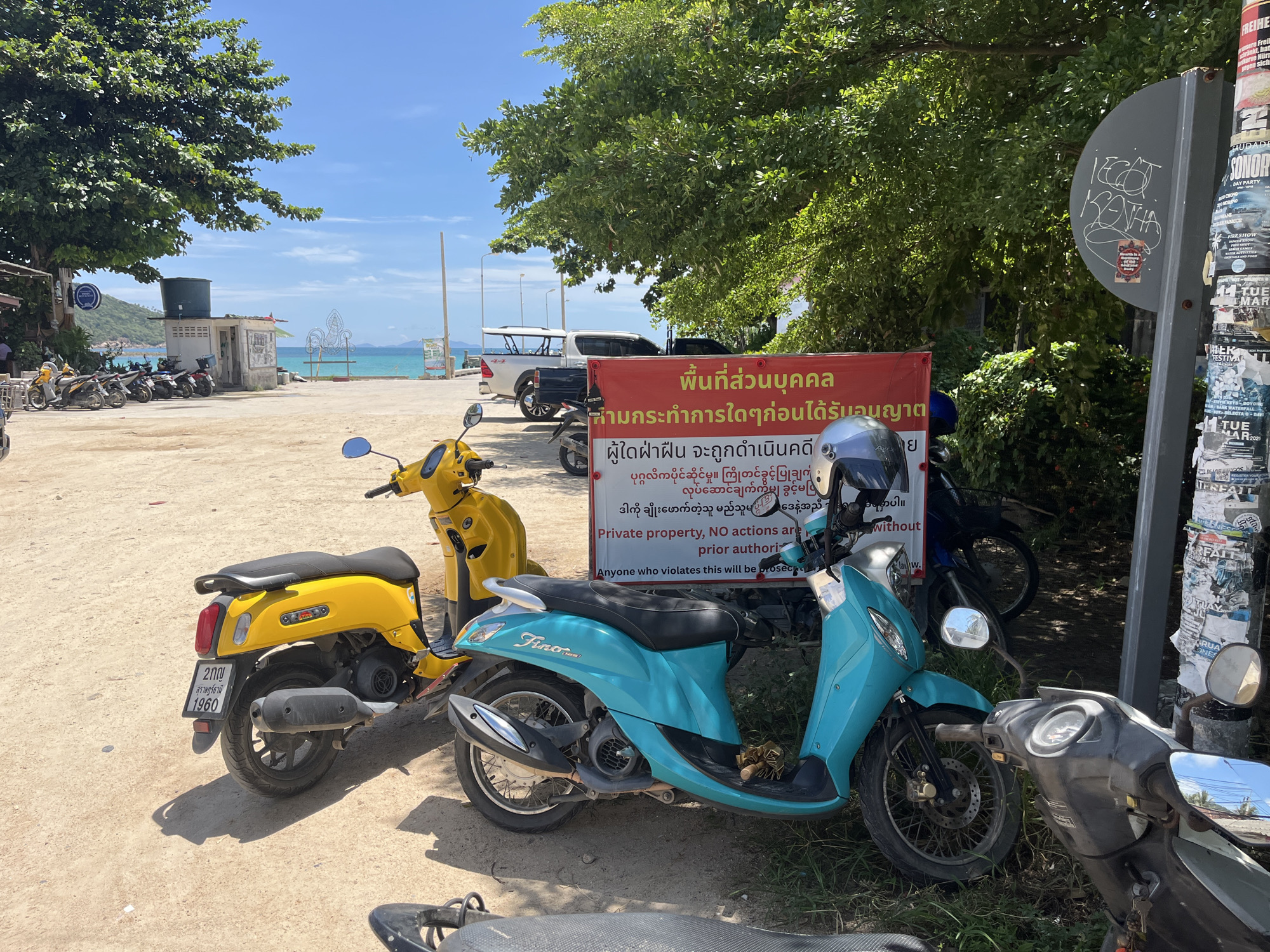 The image depicts a roadside scene in ตำบลเกาะพะงัน, Thailand. In the foreground, two scooters are parked on a dirt and gravel surface. A bright yellow scooter is on the left, and a turquoise scooter is to its right. Behind them, another scooter is partially visible. A red banner with text in Thai and English is displayed prominently, attached to a pole. The English text on the banner reads "Private property, NO actions are prior authorization. Anyone who violates this will be prosecuted." To the right of the banner, a metal pole is covered with various posters and flyers. Lush green trees provide shade over the scene. In the background, a glimpse of the ocean and sky can be seen, suggesting a coastal location. The overall impression is of a sunny day in a tropical environment.