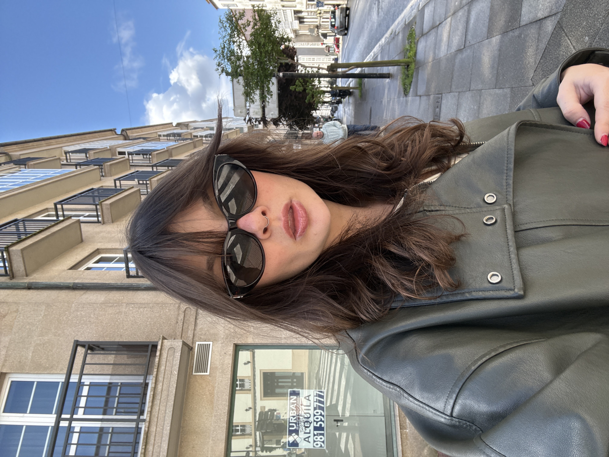A person with long brown hair, wearing sunglasses and a green leather jacket, is standing on a sidewalk in Santiago de Compostela, Spain. The person is facing the camera, with buildings visible in the background. There is a sign in a storefront window, displaying the words "URBAN ALQUILA" and the phone number "981 599 777". A street with trees and cars is visible in the distance.